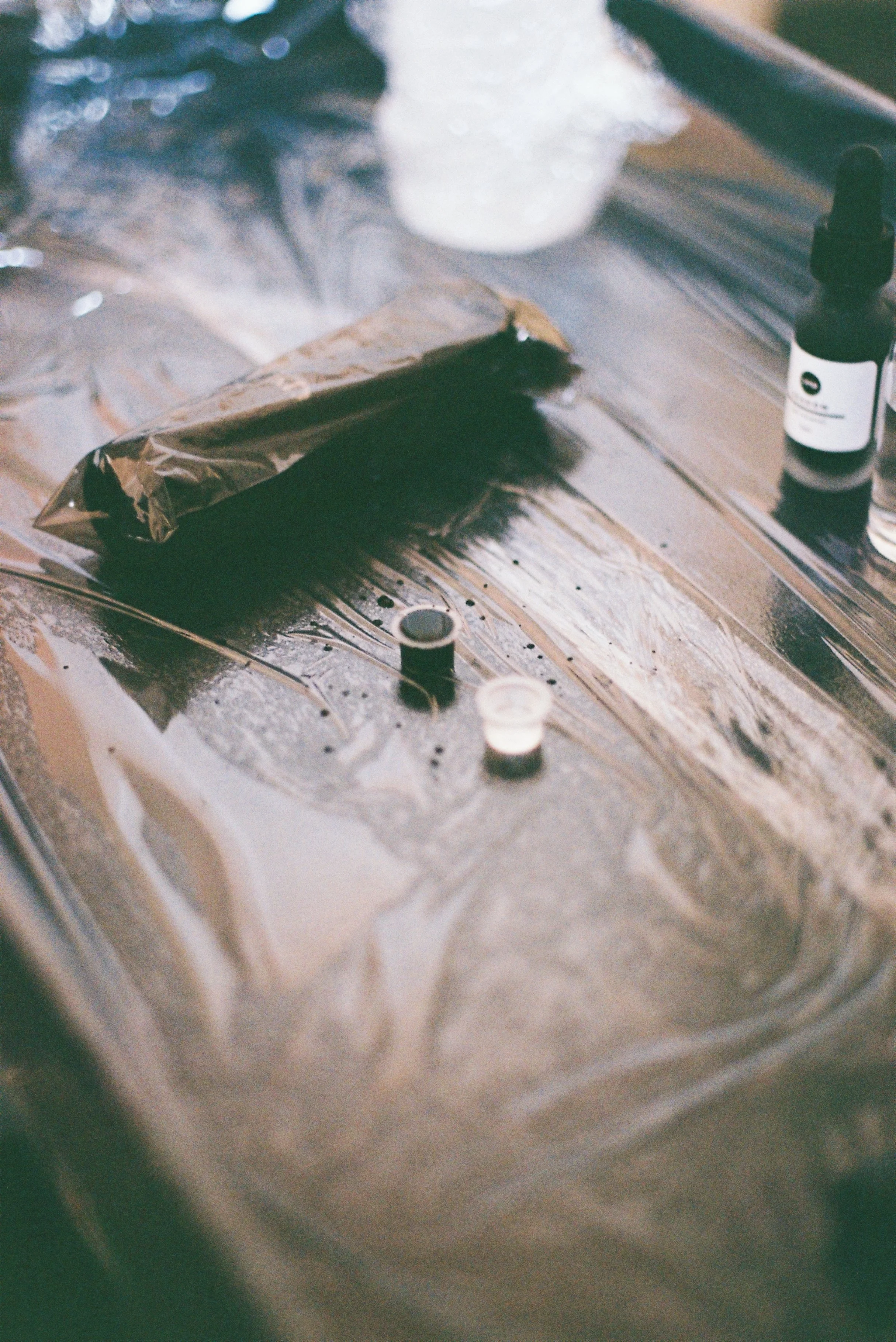 A small amber glass dropper bottle next to a black and white dropper bottle on a reflective surface, with a blurry background.