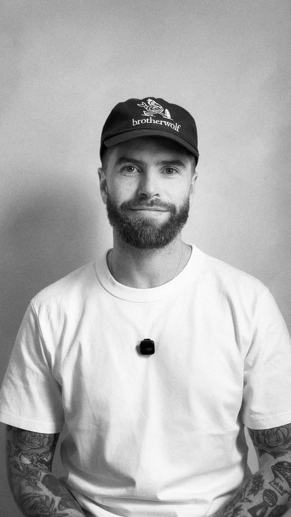 A man with a beard and tattoos on both arms, wearing a white t-shirt, a black cap with the word 'brotherwolf' and a wolf logo, and a small camera hanging around his neck, standing against a plain light-colored wall.