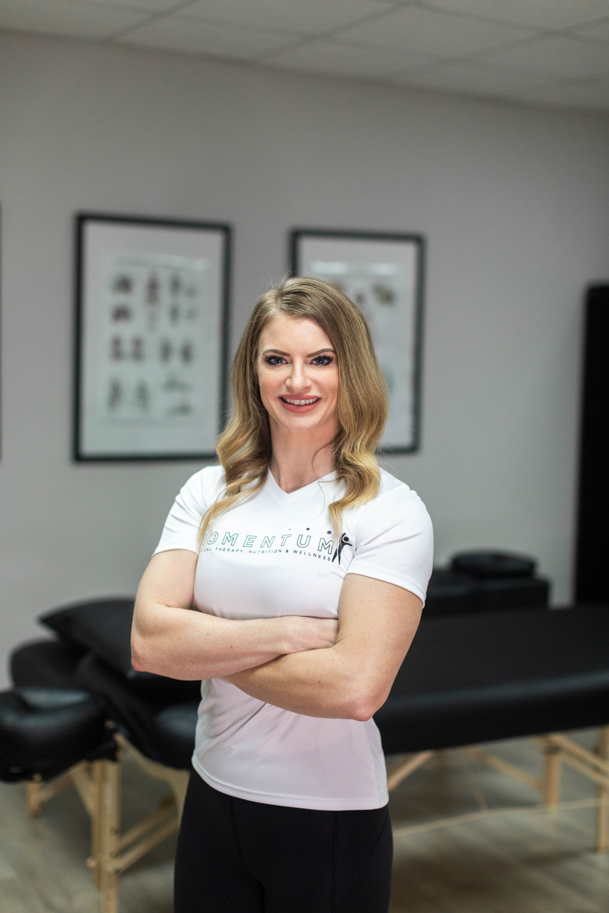 A smiling woman with long, wavy, blonde hair standing with arms crossed in a wellness clinic, in front of a massage table and framed wellness posters on the wall.
