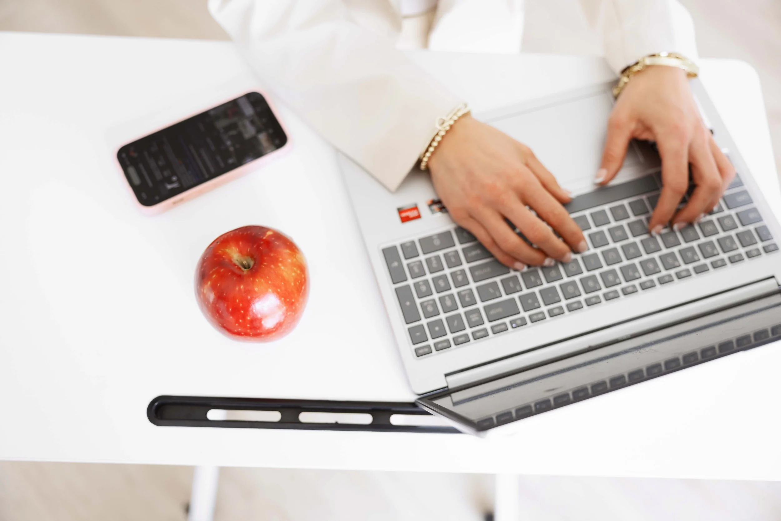 Person typing on a silver laptop at a white desk, with a red apple, a pink smartphone, and a black ruler nearby.