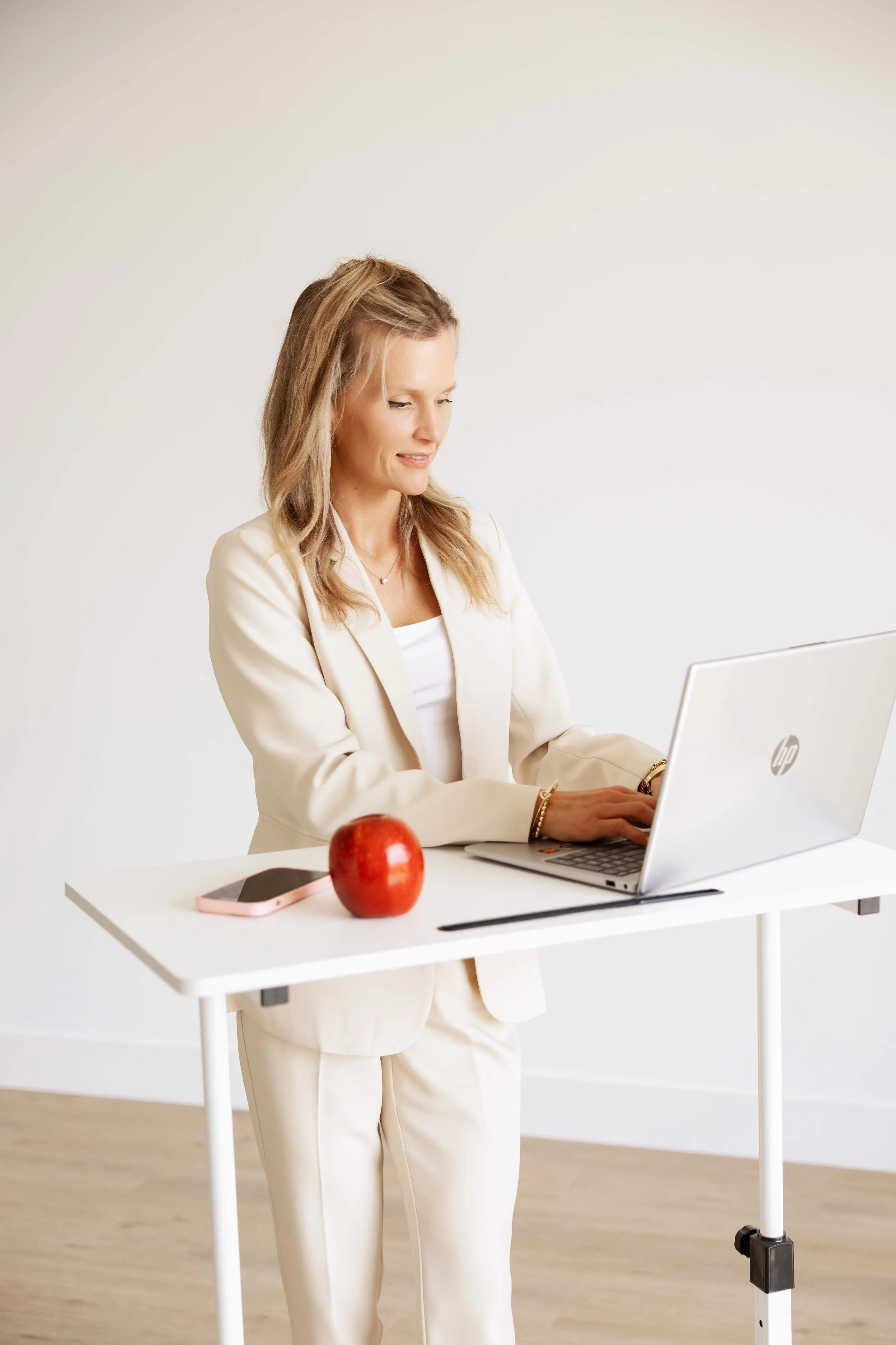 A woman in a light-colored suit working on a silver HP laptop at a white desk. There is a red apple and a smartphone on the desk. The background is plain white.