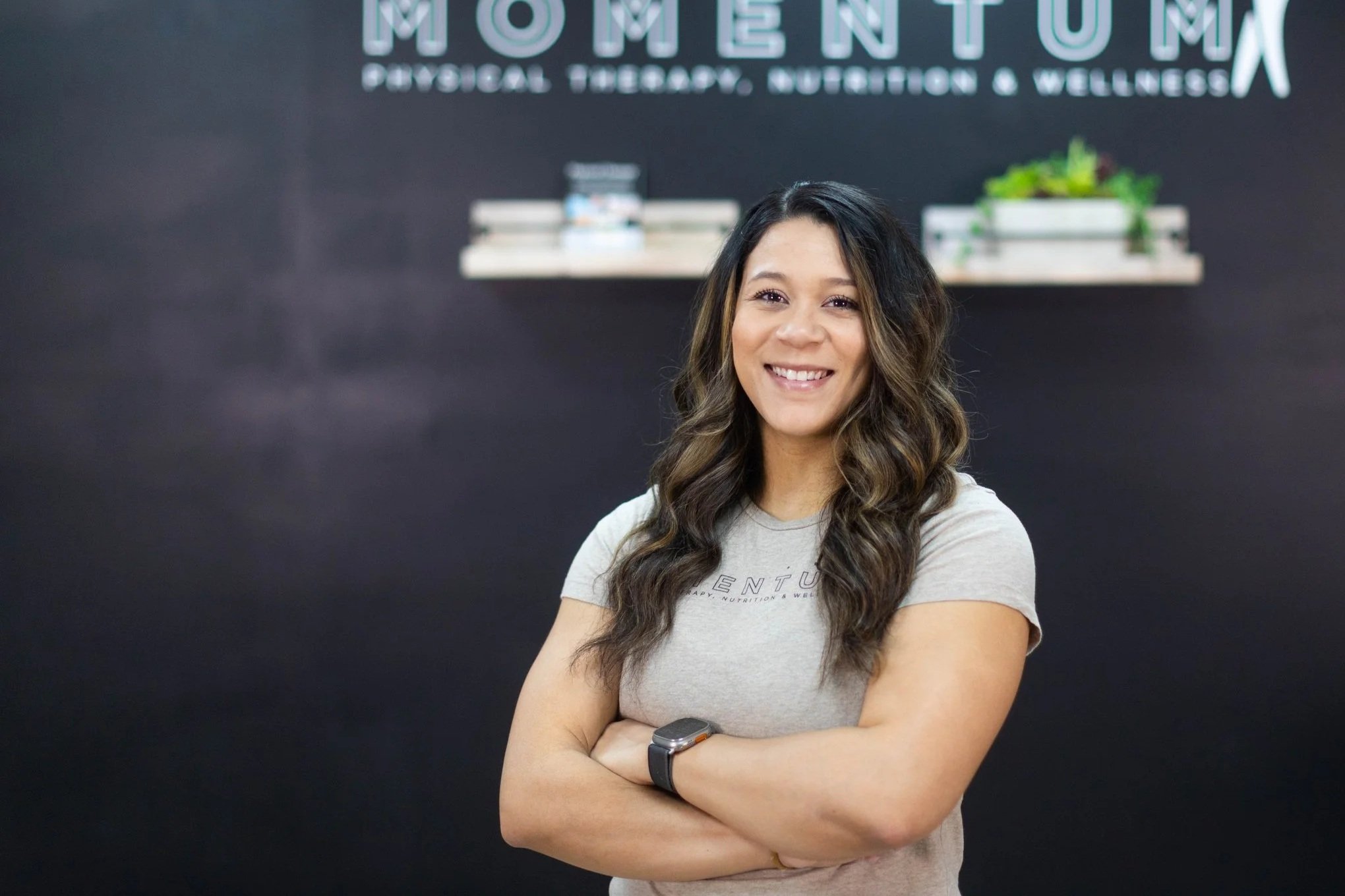 A woman with long, wavy dark brown hair, smiling with arms crossed, standing in front of a wall with logo text for Momentum Physical Therapy, Nutrition & Wellness.