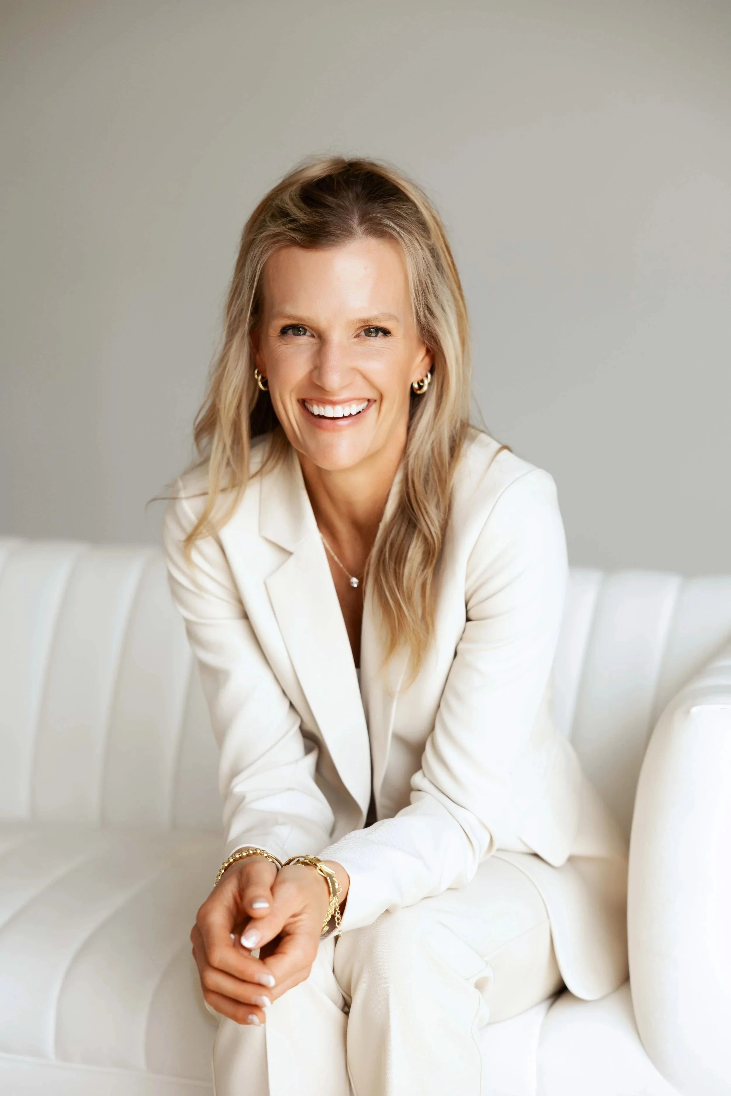 A woman with blonde hair smiling, dressed in an all-white suit, sitting on a white sofa in front of a plain gray wall.