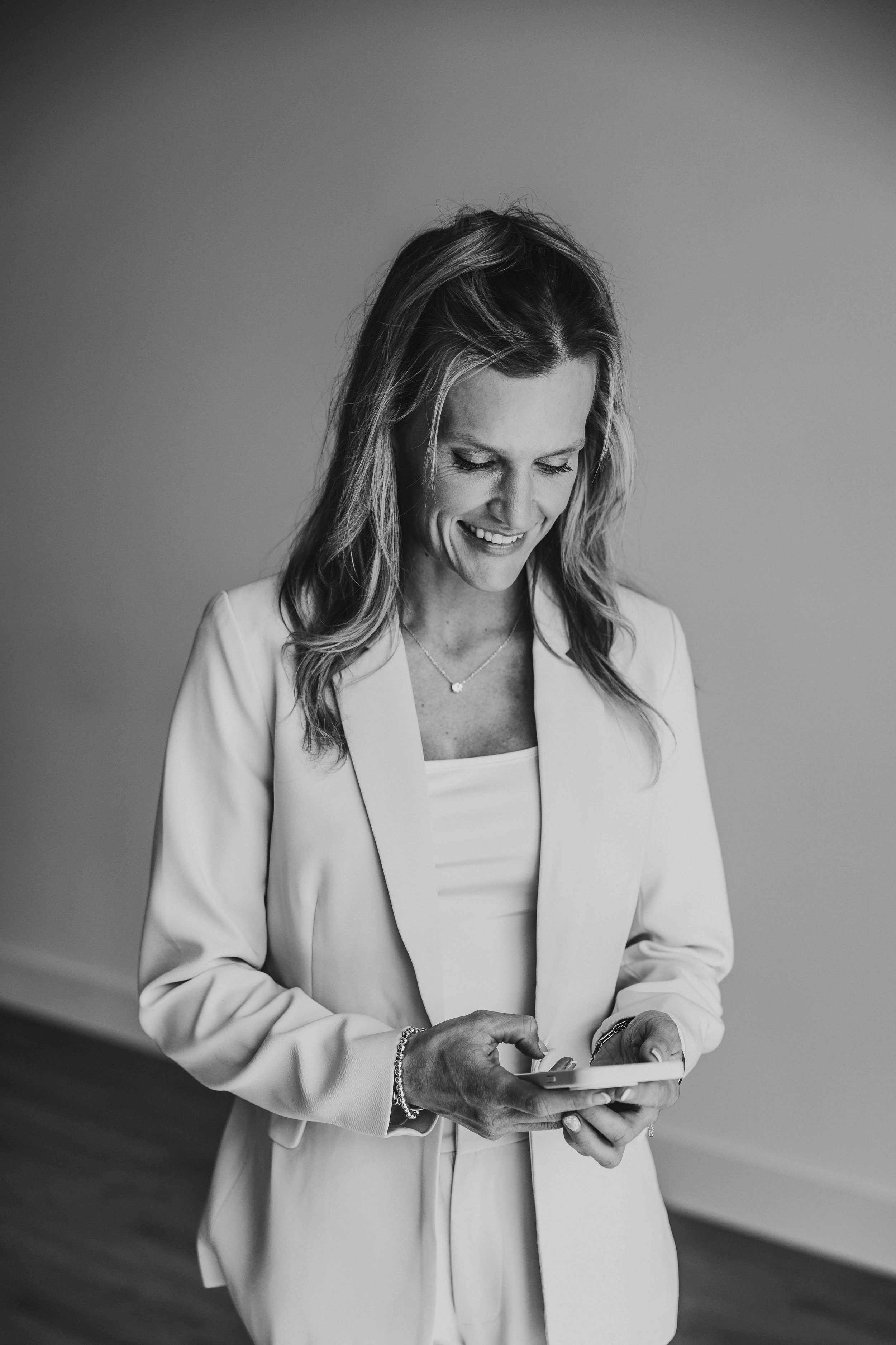 Black and white photo of a woman smiling and looking down at her phone, dressed in a business suit.