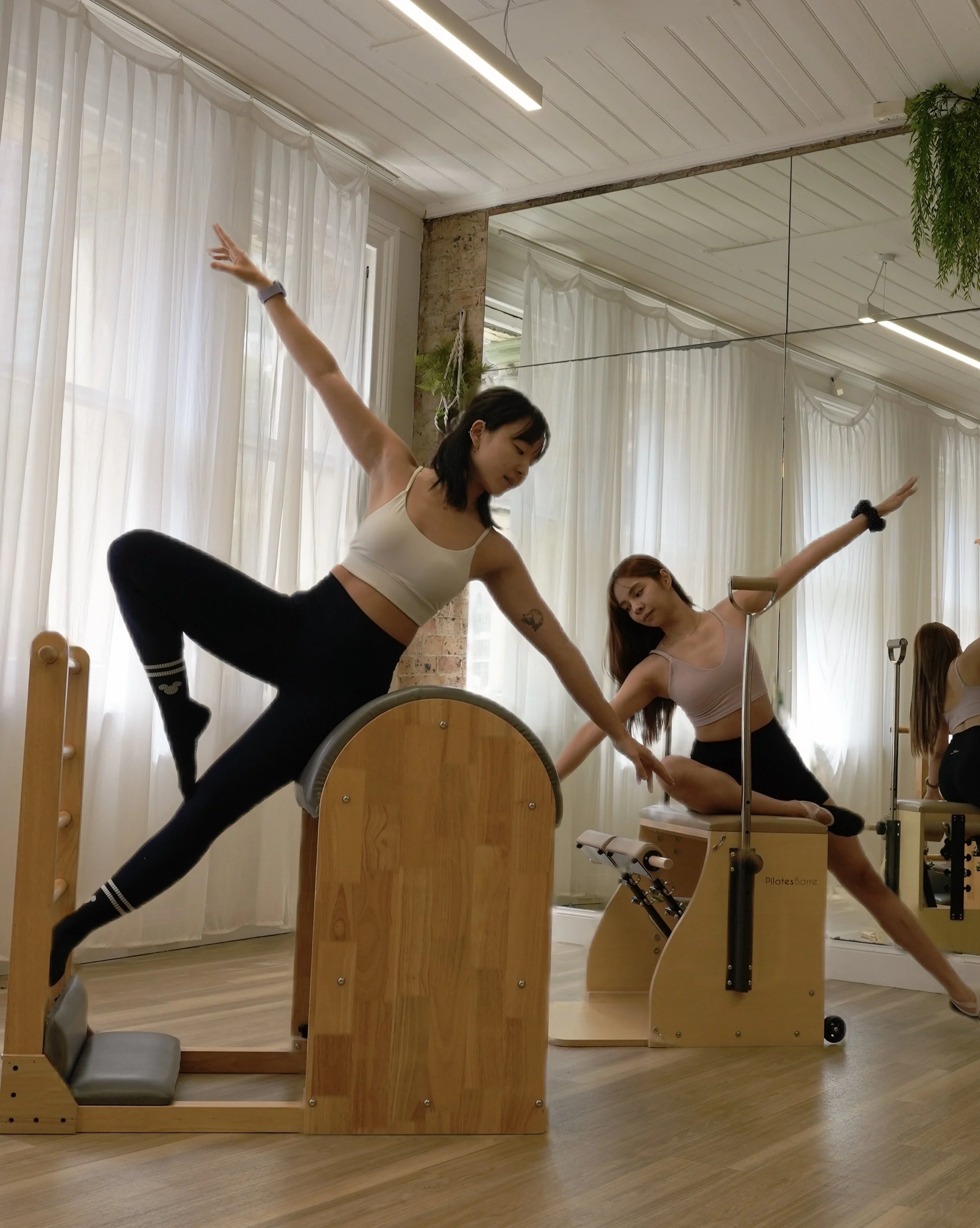 Two women practicing ballet stretches in a dance studio with large windows, white curtains, and wooden flooring, using Pilates equipment.