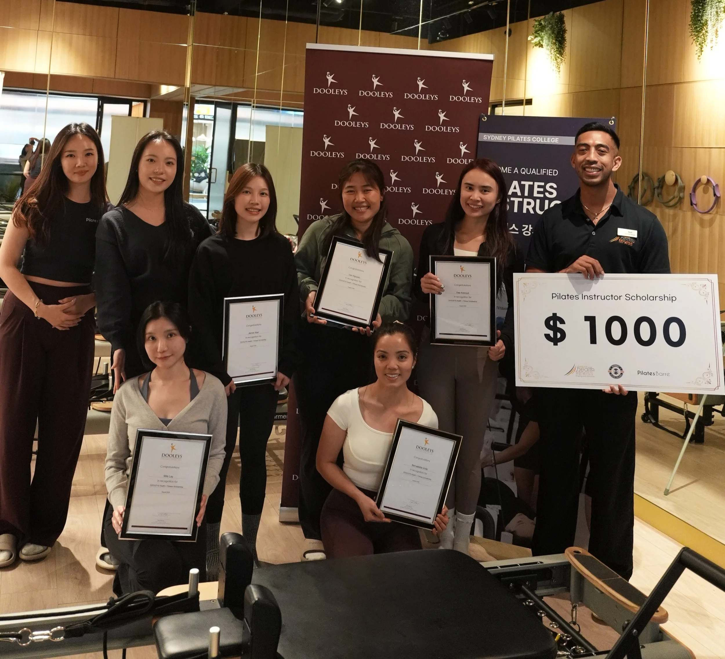 Group of six individuals holding framed certificates, standing in front of banners with logos for Dooleys and Sydney Pilates College in a room with wood-paneled walls and plants.