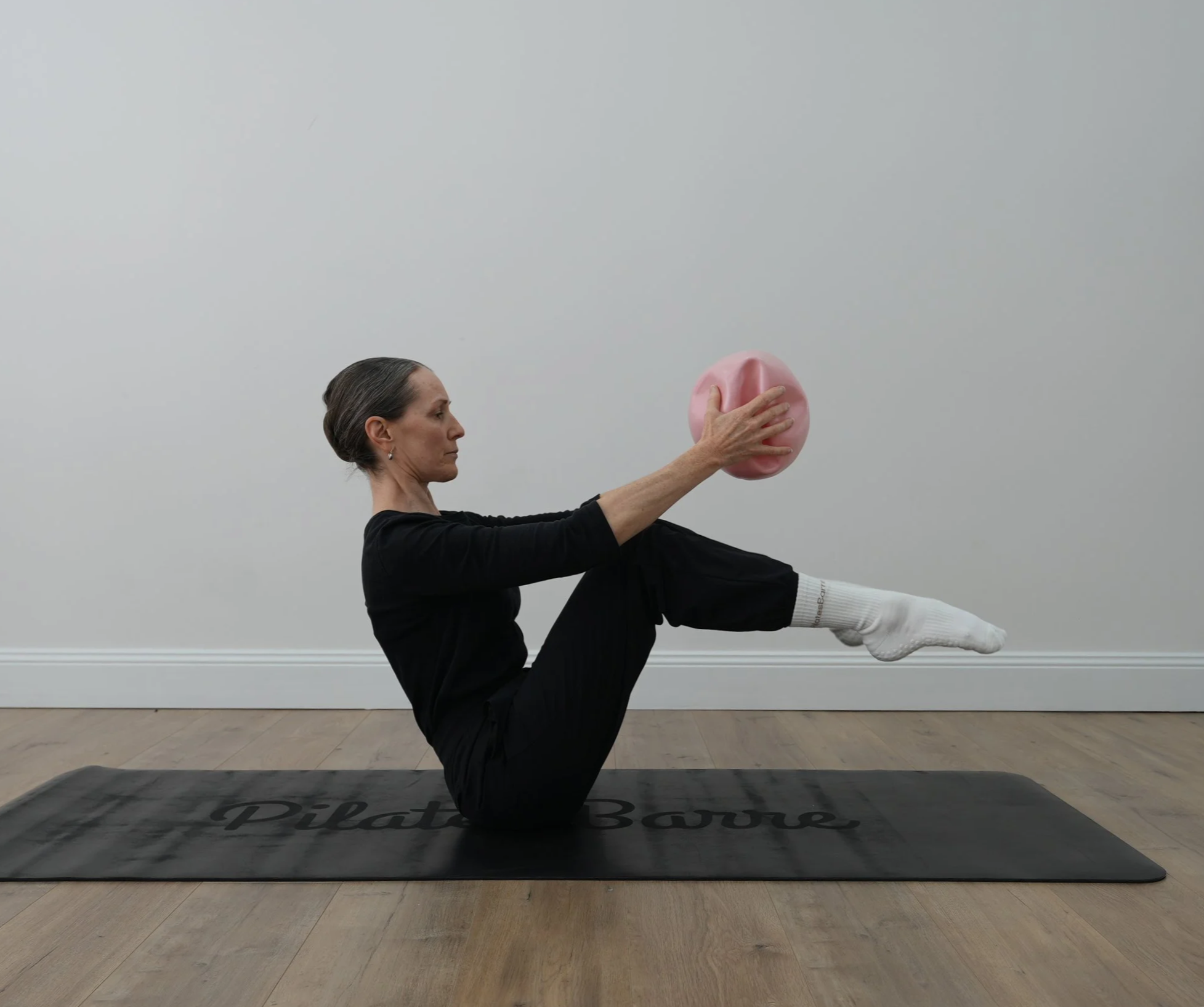 A woman in black clothing doing a seated yoga pose on a black mat with 'Pilates Body' written on it. She is holding a pink exercise ball with her right hand. The setting appears to be an indoor studio with a plain white wall and hardwood floor.