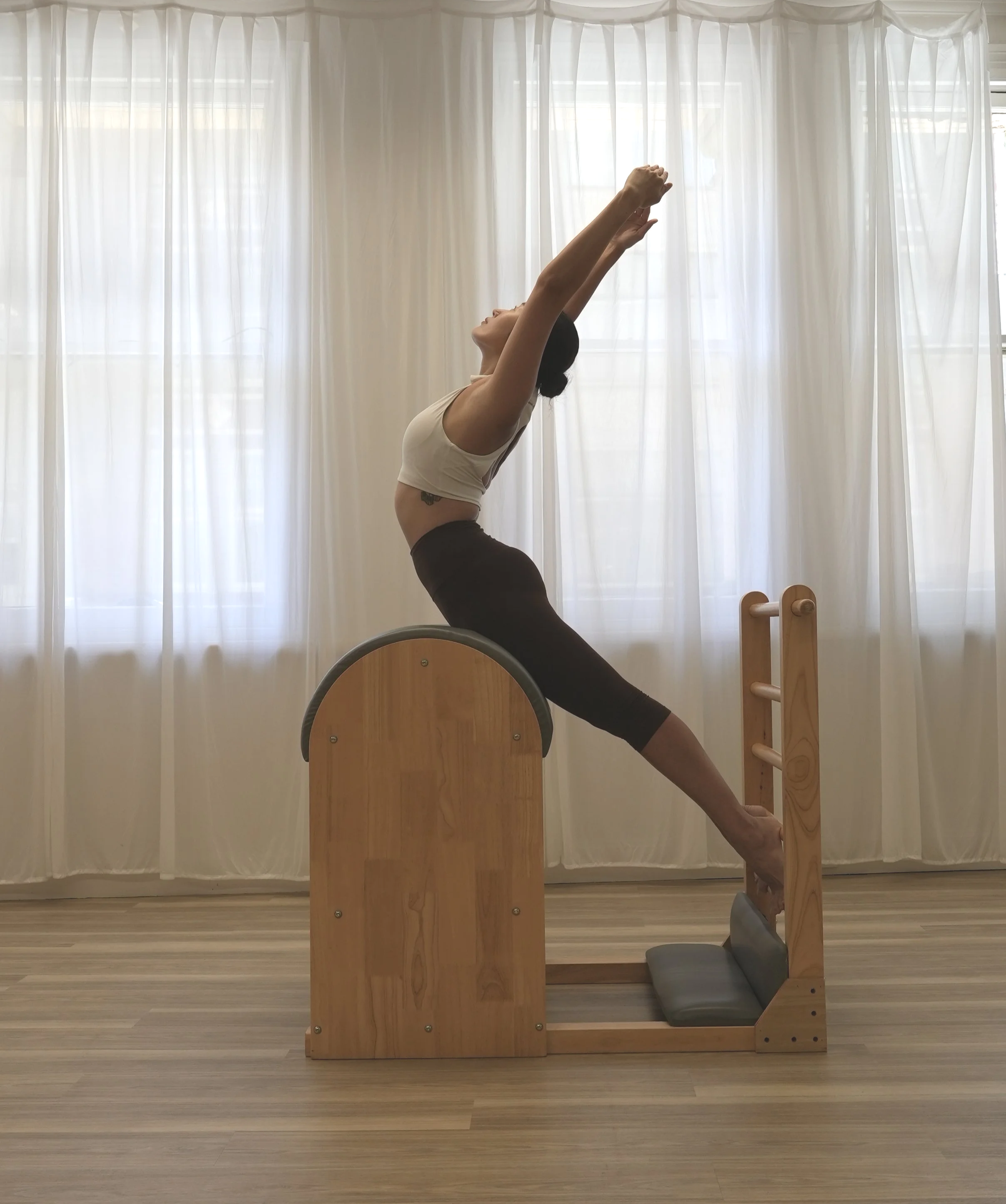 A woman practicing yoga or stretching indoors near a large window with white curtains. She is in a lunge position with her arms stretched upward, wearing a beige tank top and black leggings. There is a wooden Pilates or yoga reformer and a padded mat on the floor.