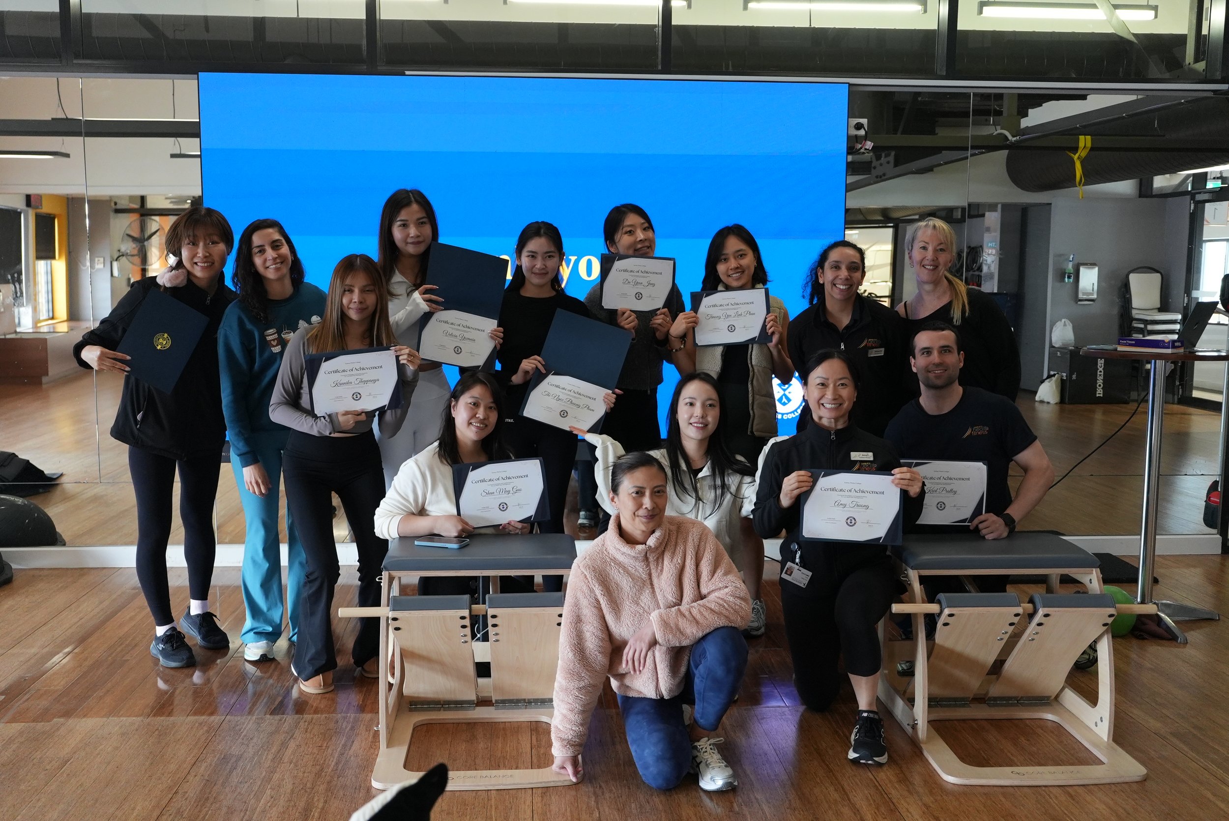 A group of diverse people at an award ceremony in a gym or fitness center, holding certificates of achievement. Some are standing, others sitting, with a large blue digital screen behind them displaying a logo or text.