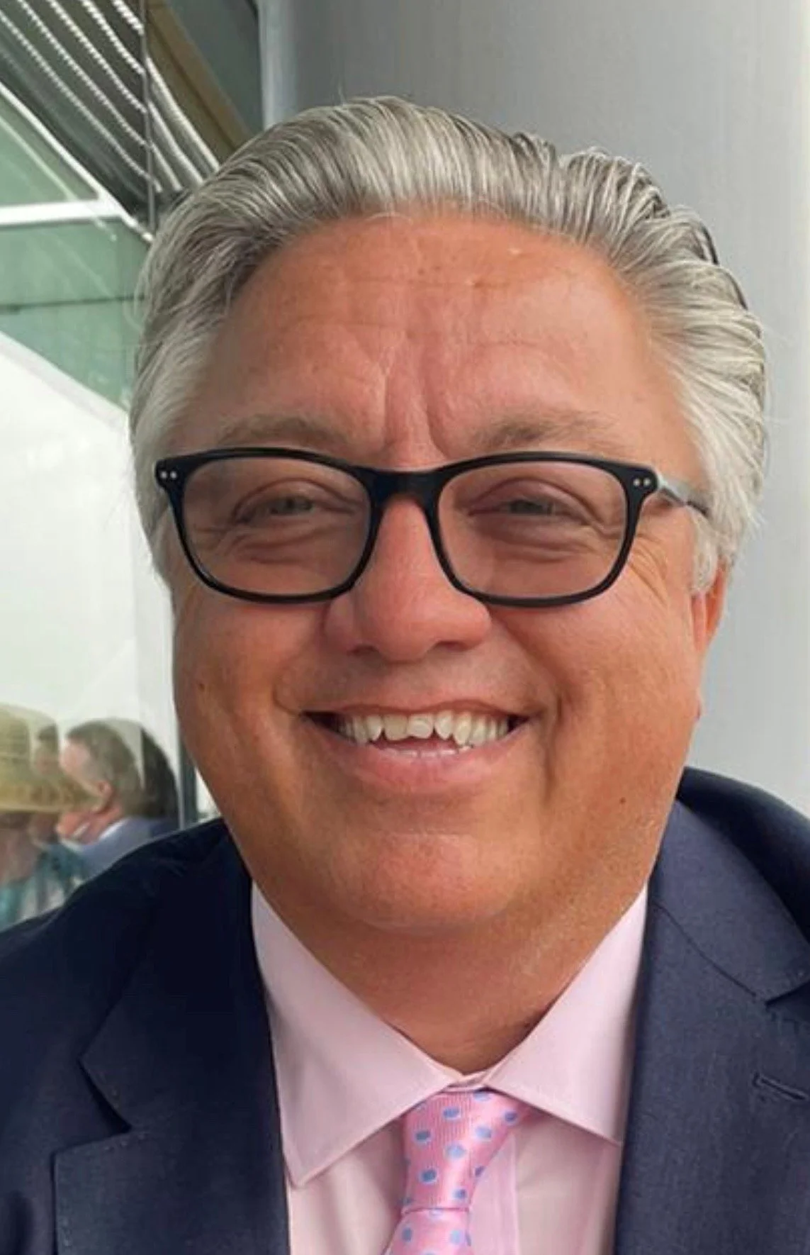 A smiling man with gray hair, black glasses, wearing a pink shirt, pink tie with blue dots, and a navy suit jacket, in an indoor setting.
