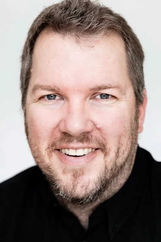 A smiling man with short, light brown hair and a beard, wearing a black shirt, against a plain light background.