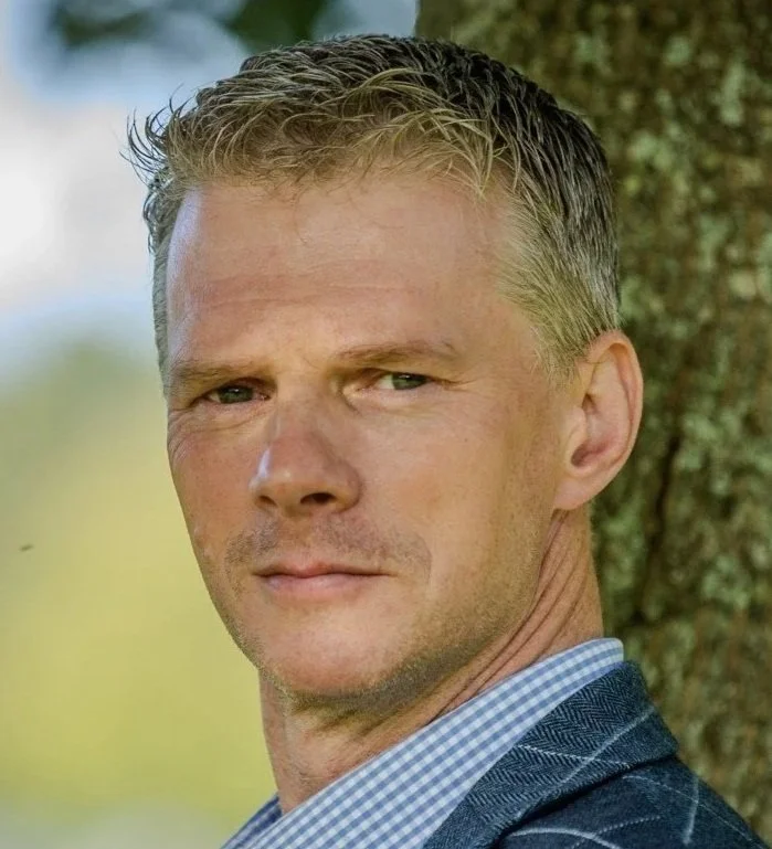 A middle-aged man with short blonde hair and blue eyes, dressed in a checkered shirt and dark jacket, standing outdoors near a tree.