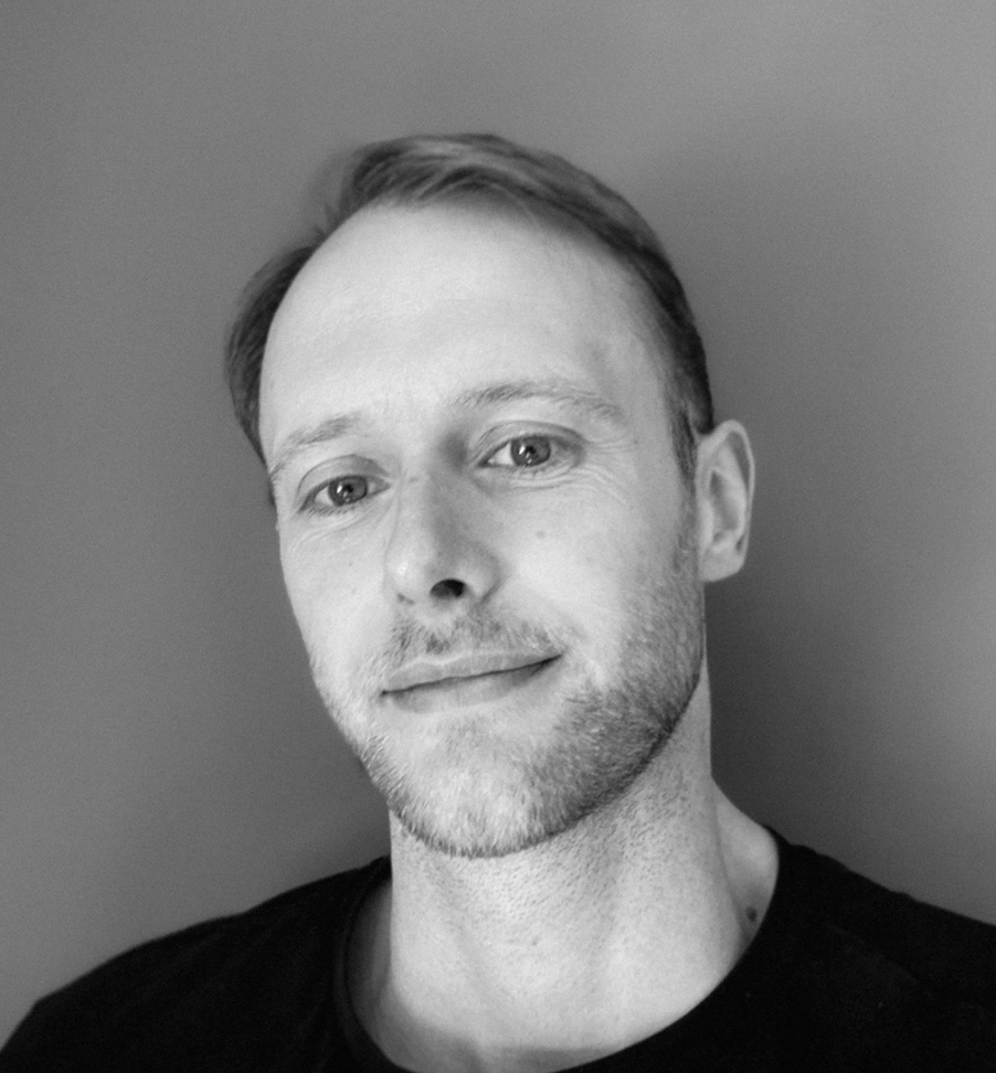 Black and white close-up portrait of a man with short hair, light facial hair, and a slight smile, wearing a dark shirt, standing against a plain background.