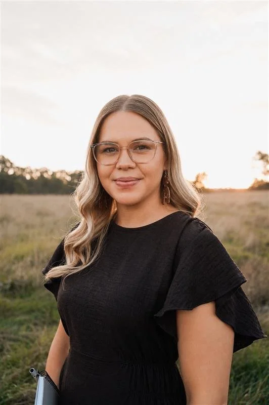 A woman with light brown, wavy hair wearing glasses and a black dress standing outdoors in a field during sunset.