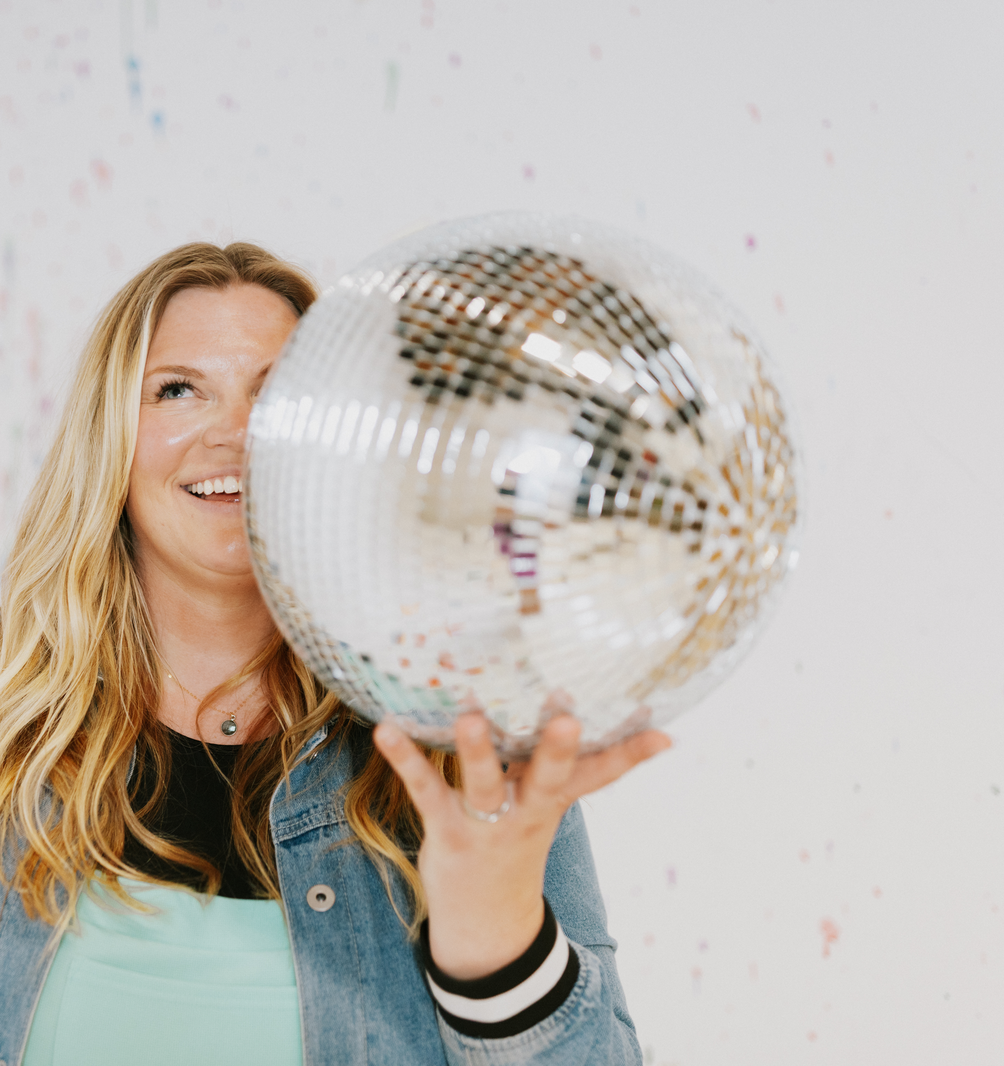 Michelle Hagen Creative. Woman with blonde hair smiling, holding a glittery disco ball in front of her face, wearing a denim jacket and light-colored top.