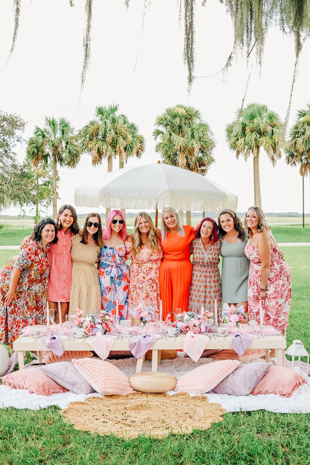 Group of eight women posing under a white patio umbrella during an outdoor gathering on a grassy field, with palm trees in the background and decorated table and seating area in front.