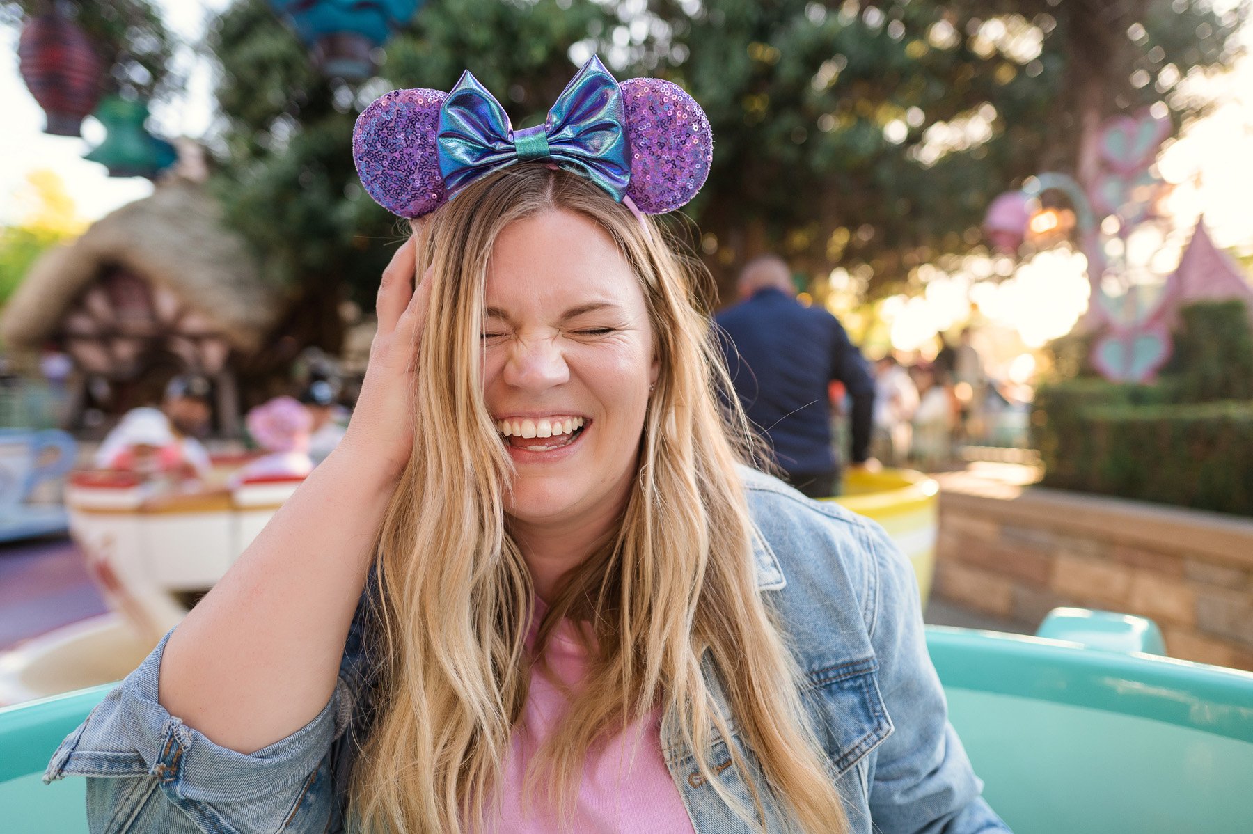 Michelle Hagen. A woman with long blonde hair wearing a purple glittery Mickey Mouse ears headband with a shiny blue bow, smiling and holding her head with one hand at Disney World.