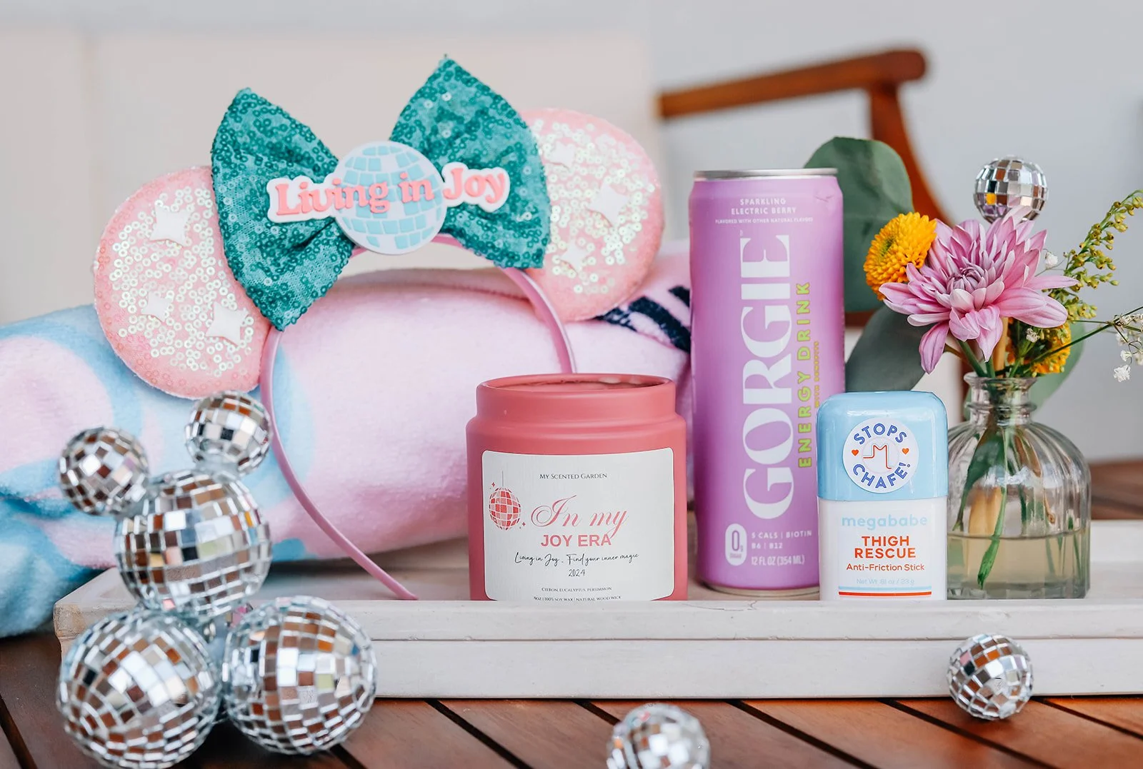 A collection of wellness and self-care items on a white wooden tray, including a pink scented candle labeled "In my Joy Era," a pink and purple energy drink can, a blue anti-friction stick labeled "Thigh Rescue," a small bouquet of pink and yellow flowers in a glass jar, and a colorful headband with a bow and the phrase "Living in Joy". There are metallic disco ball decorations scattered around the tray.