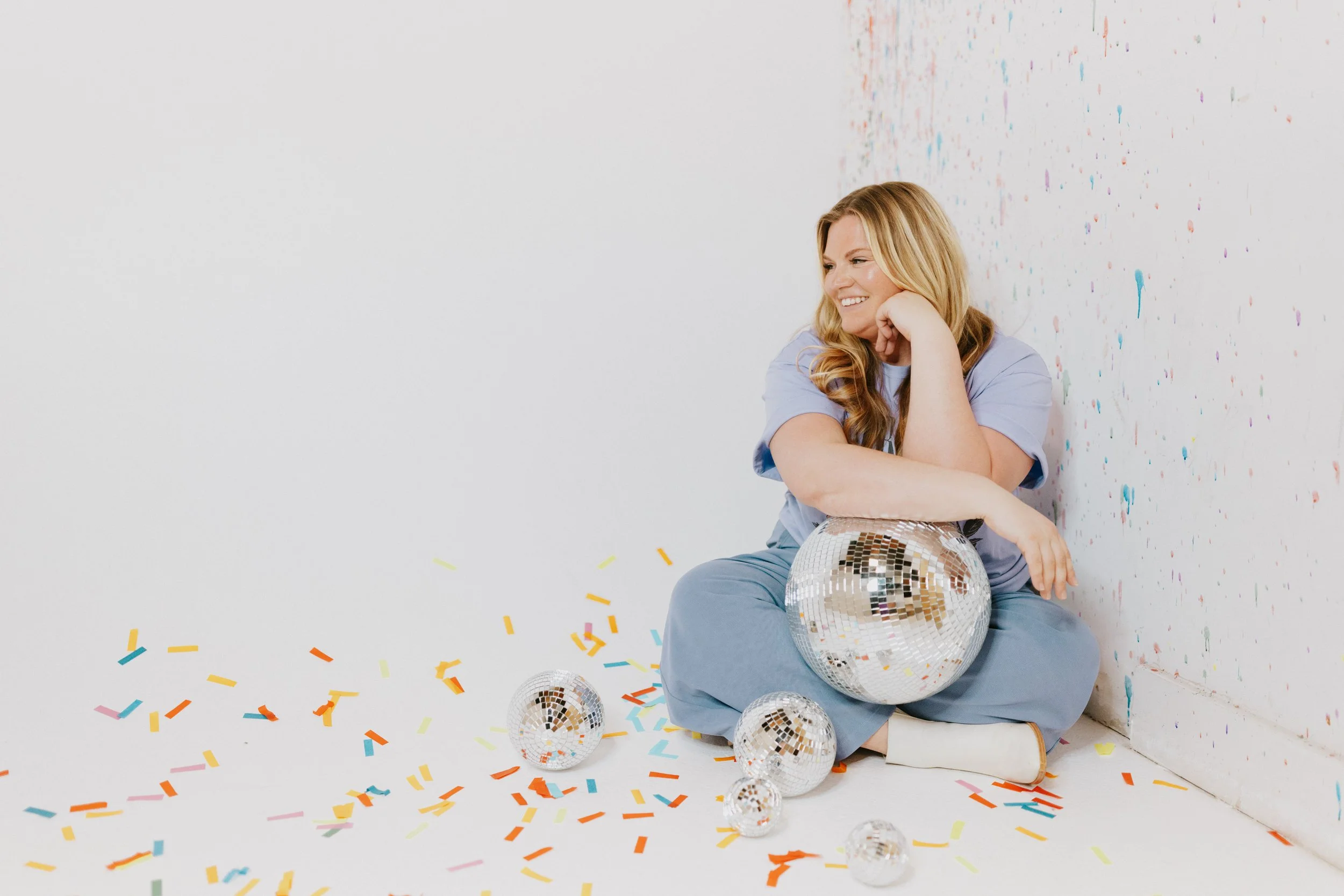 Michelle Hagen Brand Creative. A smiling woman with blonde hair wearing a blue outfit, sitting on the floor surrounded by colorful confetti and disco balls, with a white background and a multicolored splatter pattern on the wall.