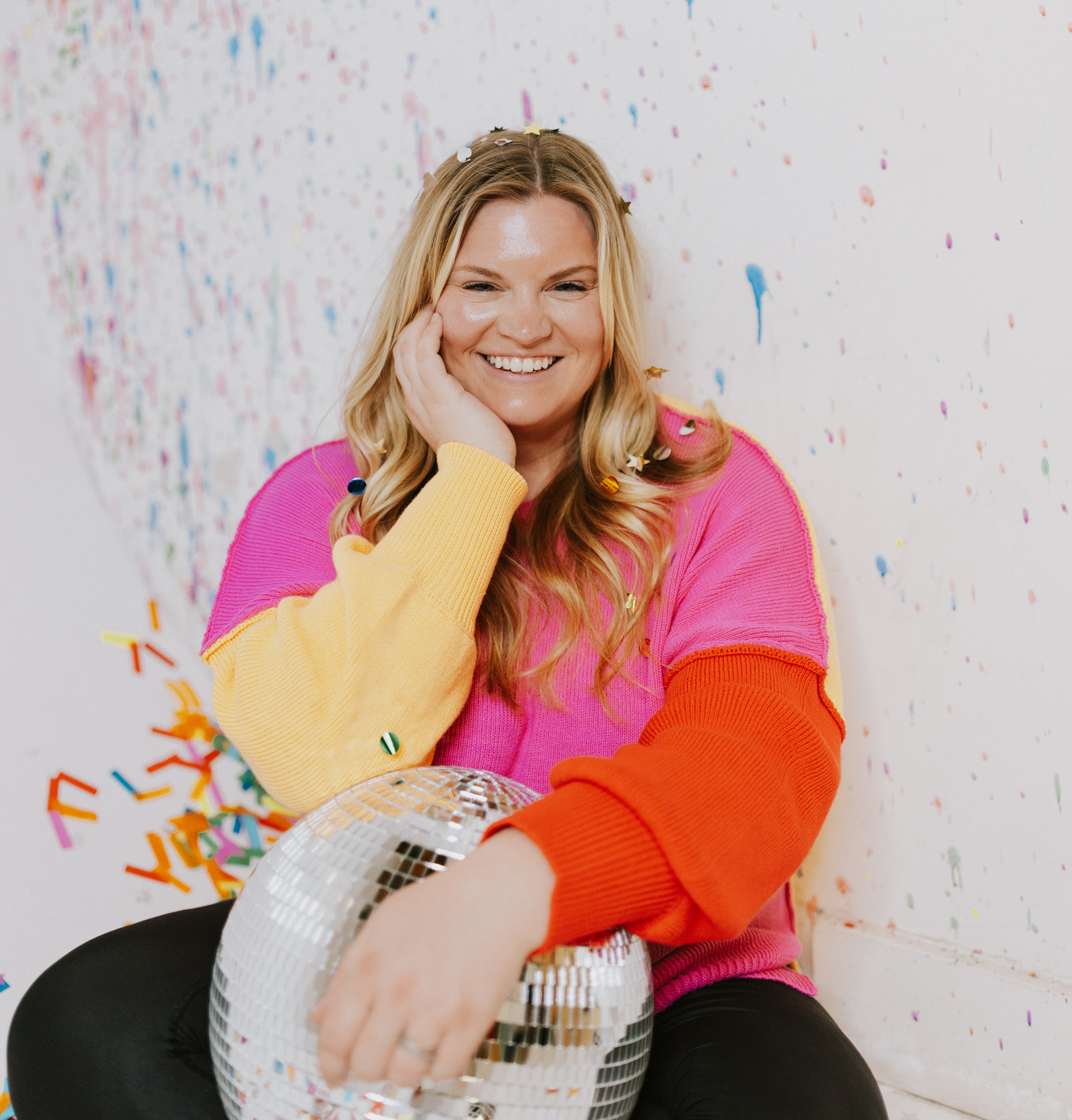 Michelle Hagen. A woman smiling, sitting against a wall with colorful confetti and streamers, wearing a pink sweater with yellow and orange accents, and a reflective disco ball in her hand.