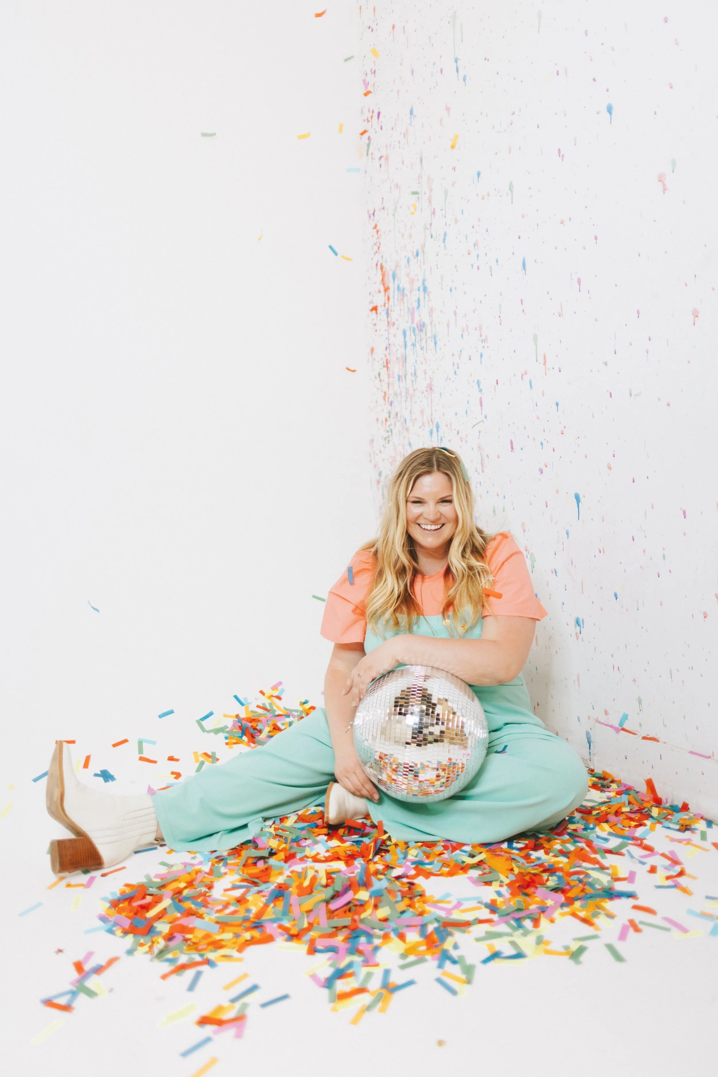Michelle Hagen. A woman sitting cross-legged on the floor surrounded by colorful confetti, holding a shiny disco ball, smiling in front of a white wall with some confetti scattered on the floor around her.