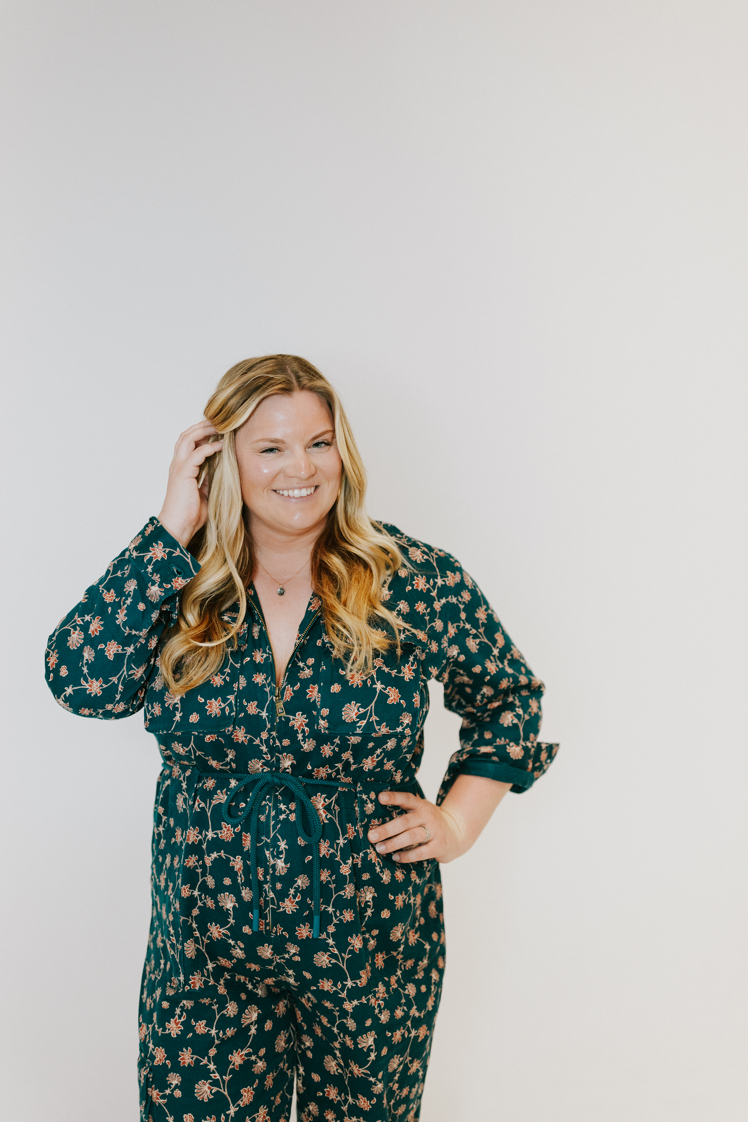 Michelle Hagen, Omaha, Nebraska. A woman with long, wavy blonde hair smiling and touching her hair, wearing a dark blue floral jumpsuit with an embroidered flower pattern, standing against a plain white background.