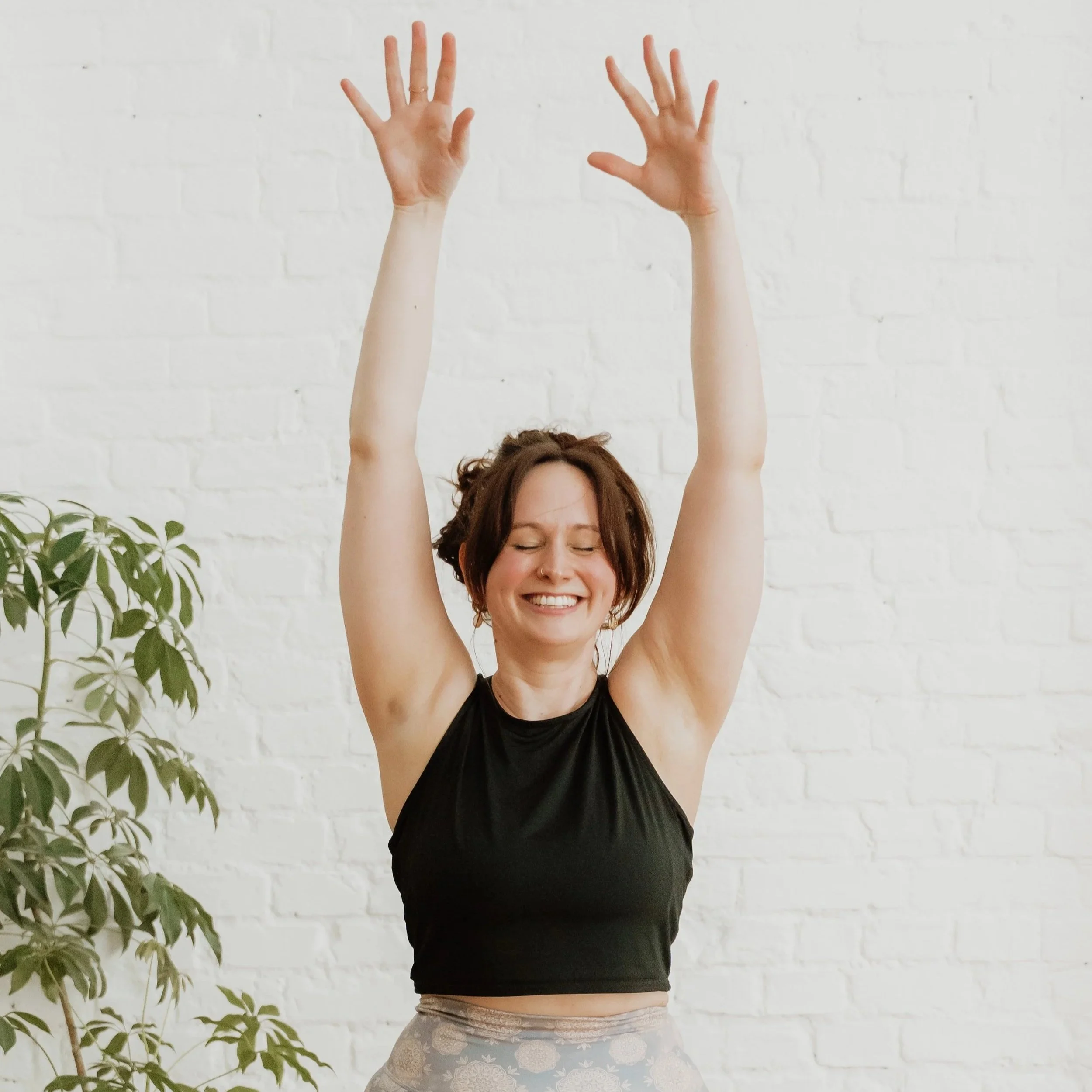 A person with curly hair smiling with eyes closed, arms raised above her head, standing against a white brick wall with a green plant on the left.