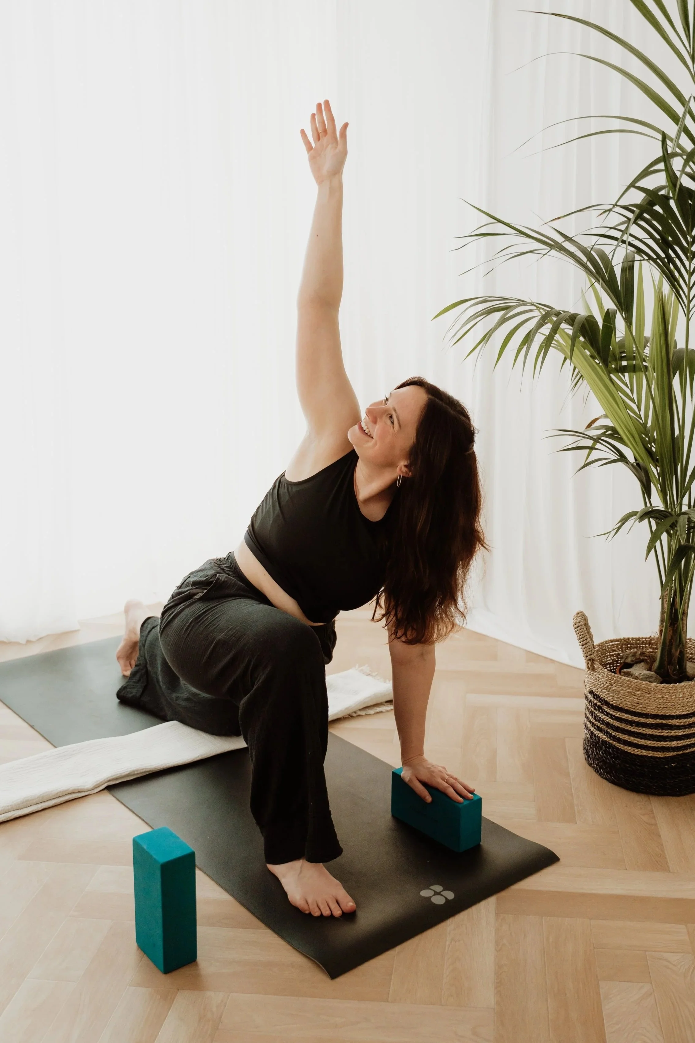 A person practicing yoga indoors, performing a twisted lunge pose with one arm raised and supported by yoga blocks, on a mat near a large plant and a basket with rocks, in a bright room with white curtains.