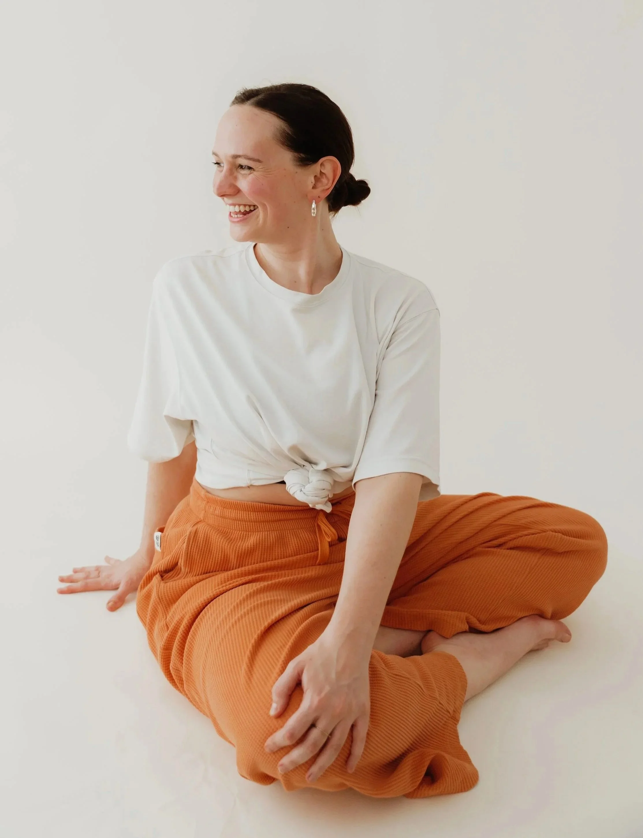 A person with dark hair tied back, wearing earrings, a white t-shirt, and orange trousers, sitting cross-legged on a white surface in a bright, minimalist setting, smiling and looking to her left.