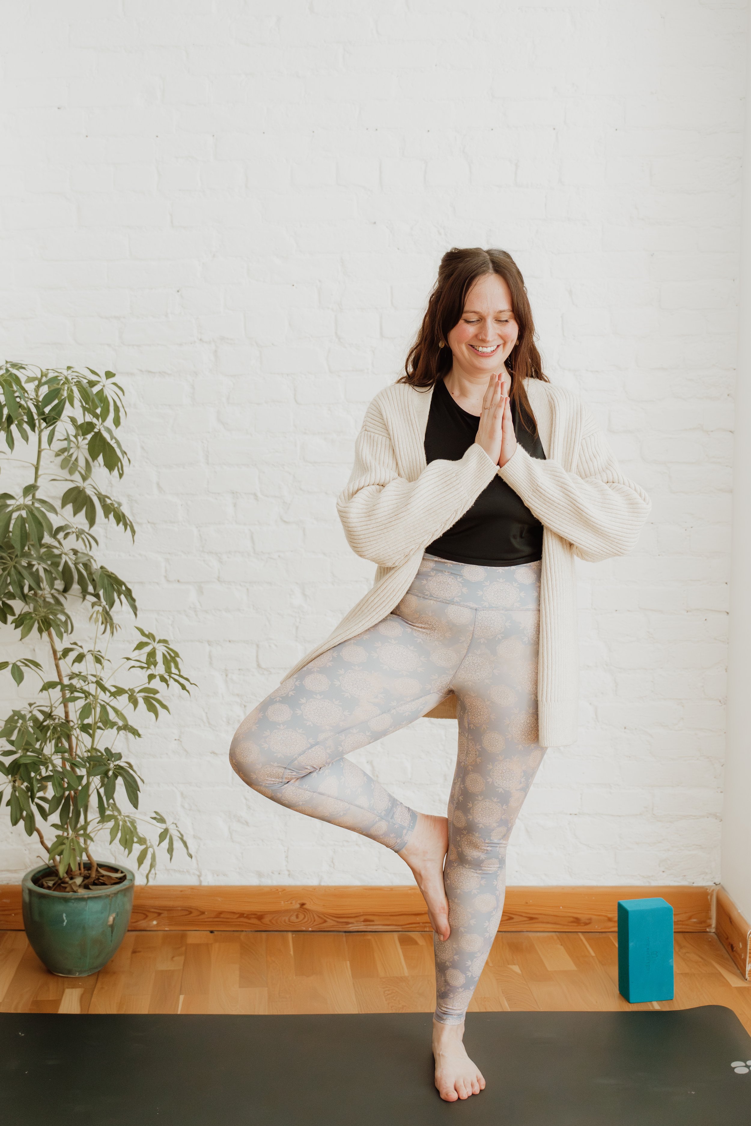 Person practicing yoga indoors in tree pose, with hands in prayer position, smiling, standing on a yoga mat with a white brick wall background, potted plant on the left, blue yoga block on the right.