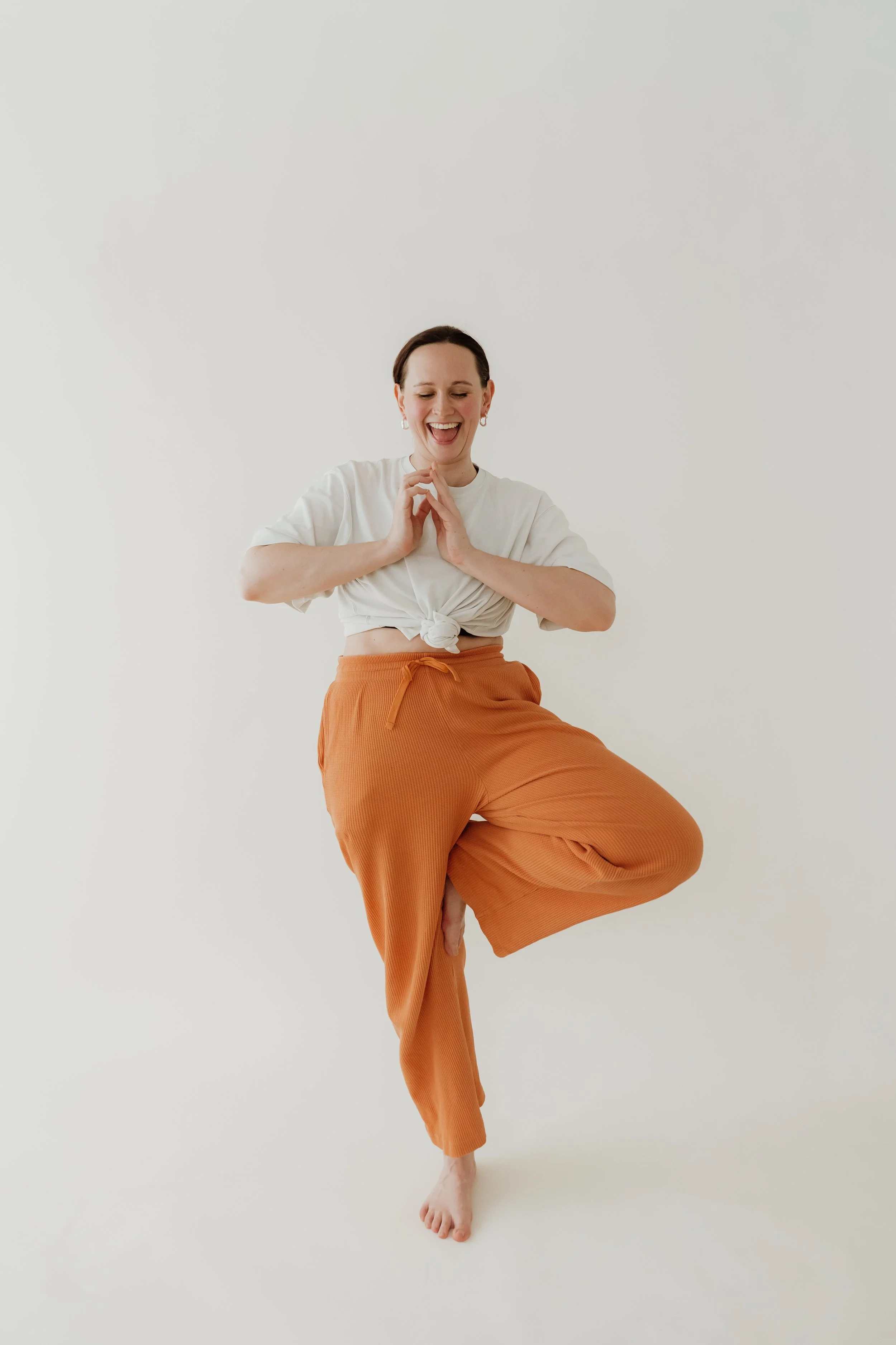 A person practicing yoga on one leg with arms in a prayer position, smiling, dressed in white knotted top and loose orange trousers, against a plain white background.