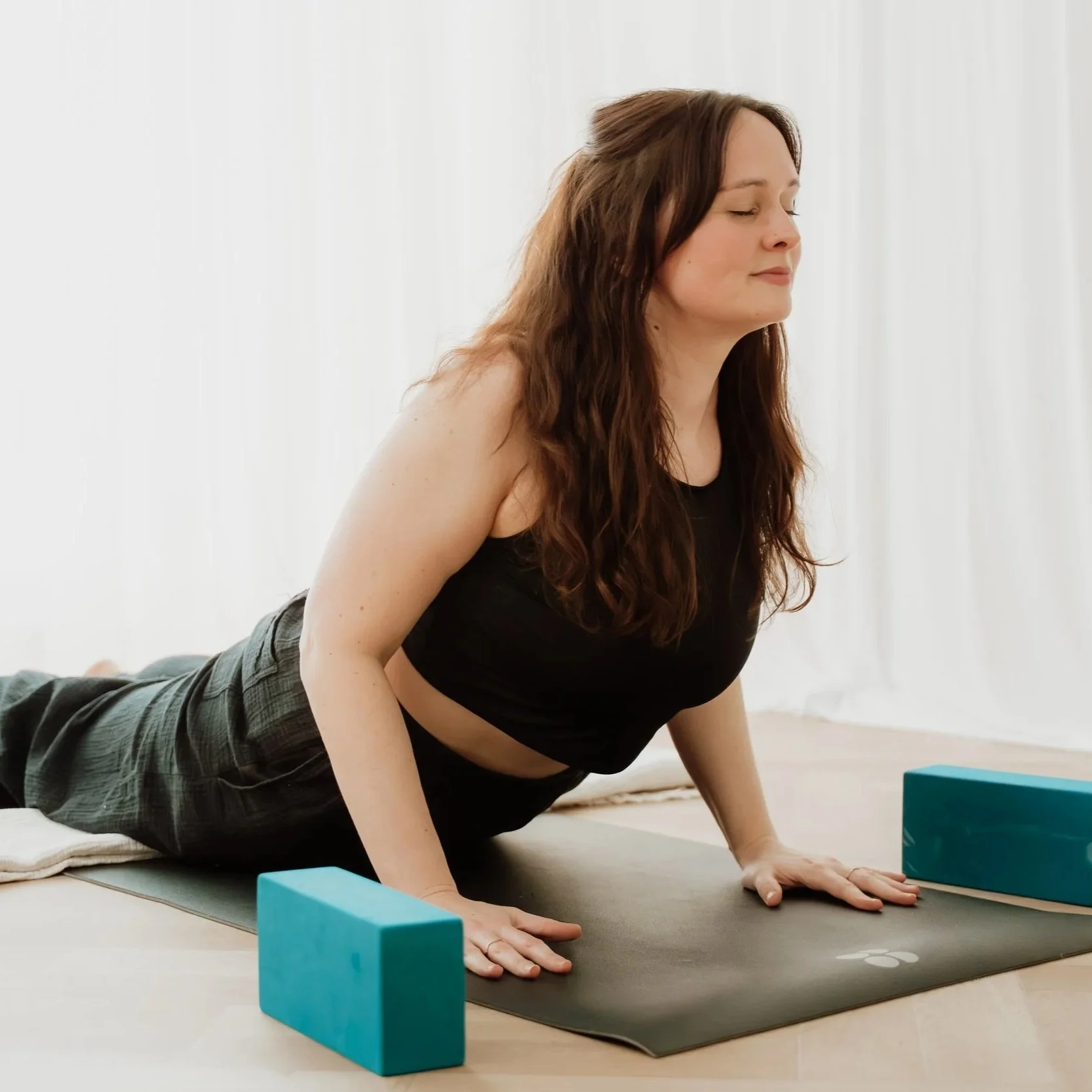 A person with long brown hair and closed eyes practicing yoga in a serene, minimalist room with natural light, using yoga blocks for support.