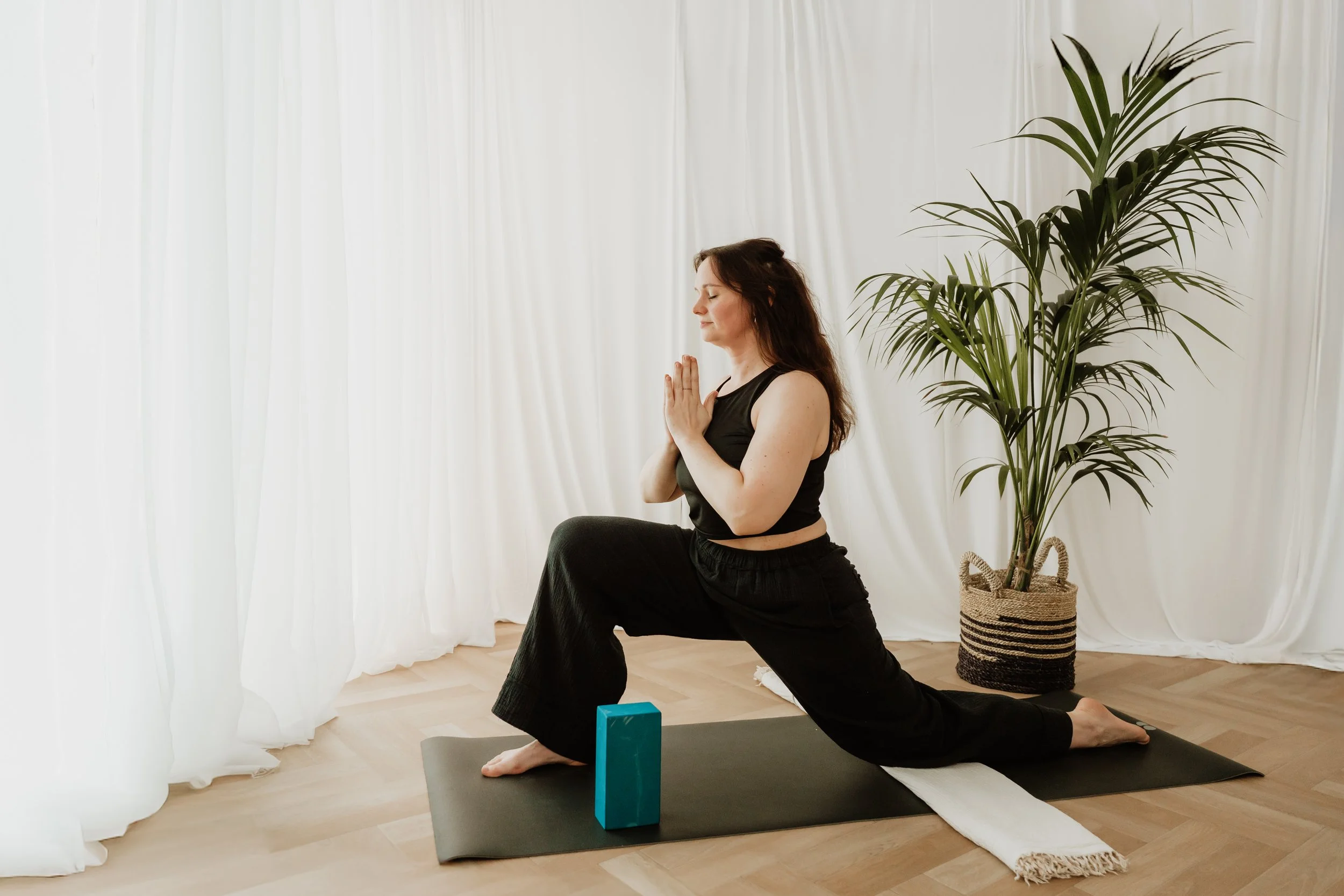 A person in loose black workout clothes practicing yoga in a bright room with white curtains, a large potted plant, and a yoga mat.