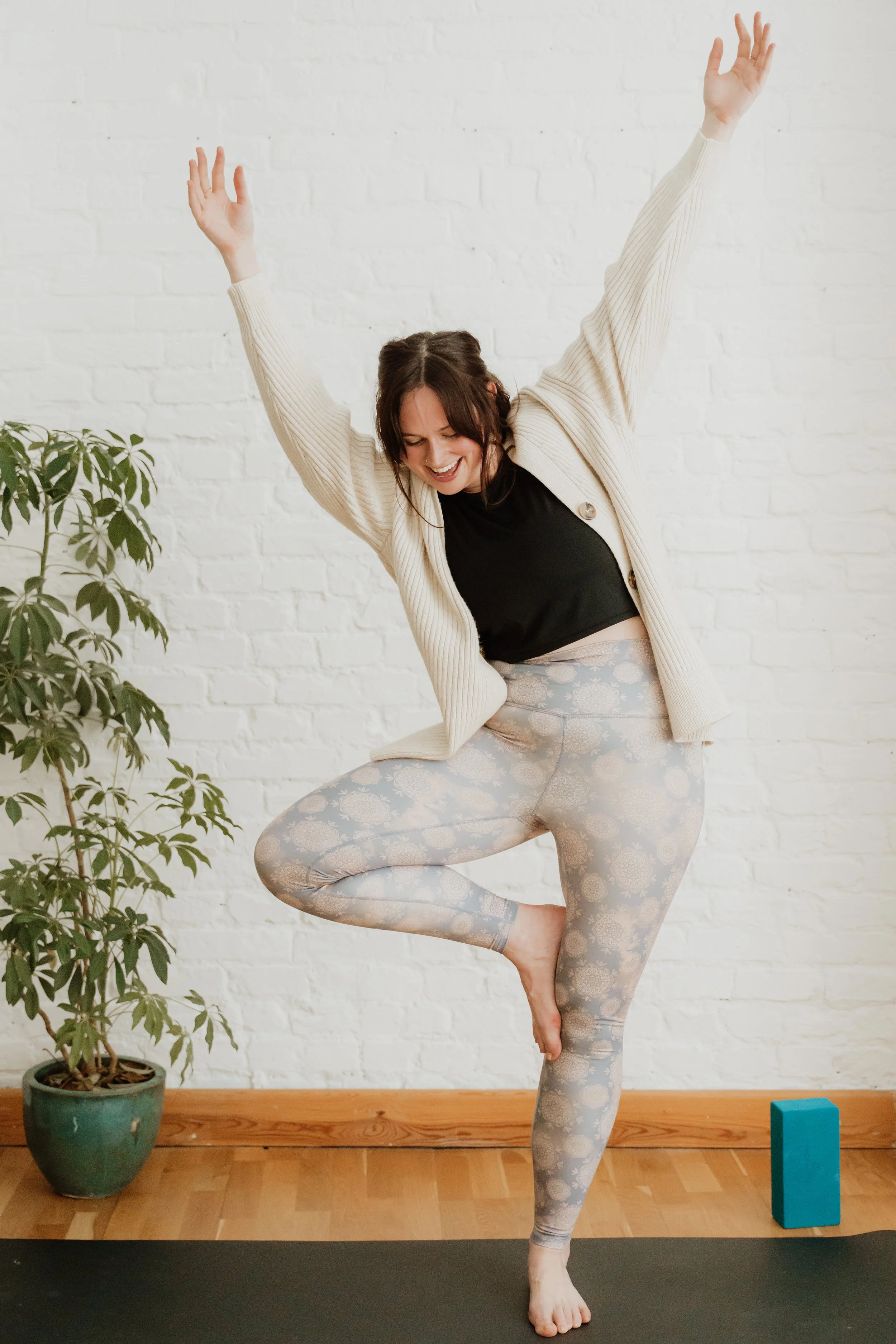 A person practicing yoga indoors, balancing on one leg, slightly wobbling, with her hands raised above her head. She is wearing patterned leggings and a black top with a cream-coloured cardigan, with a potted plant and a blue yoga block nearby.