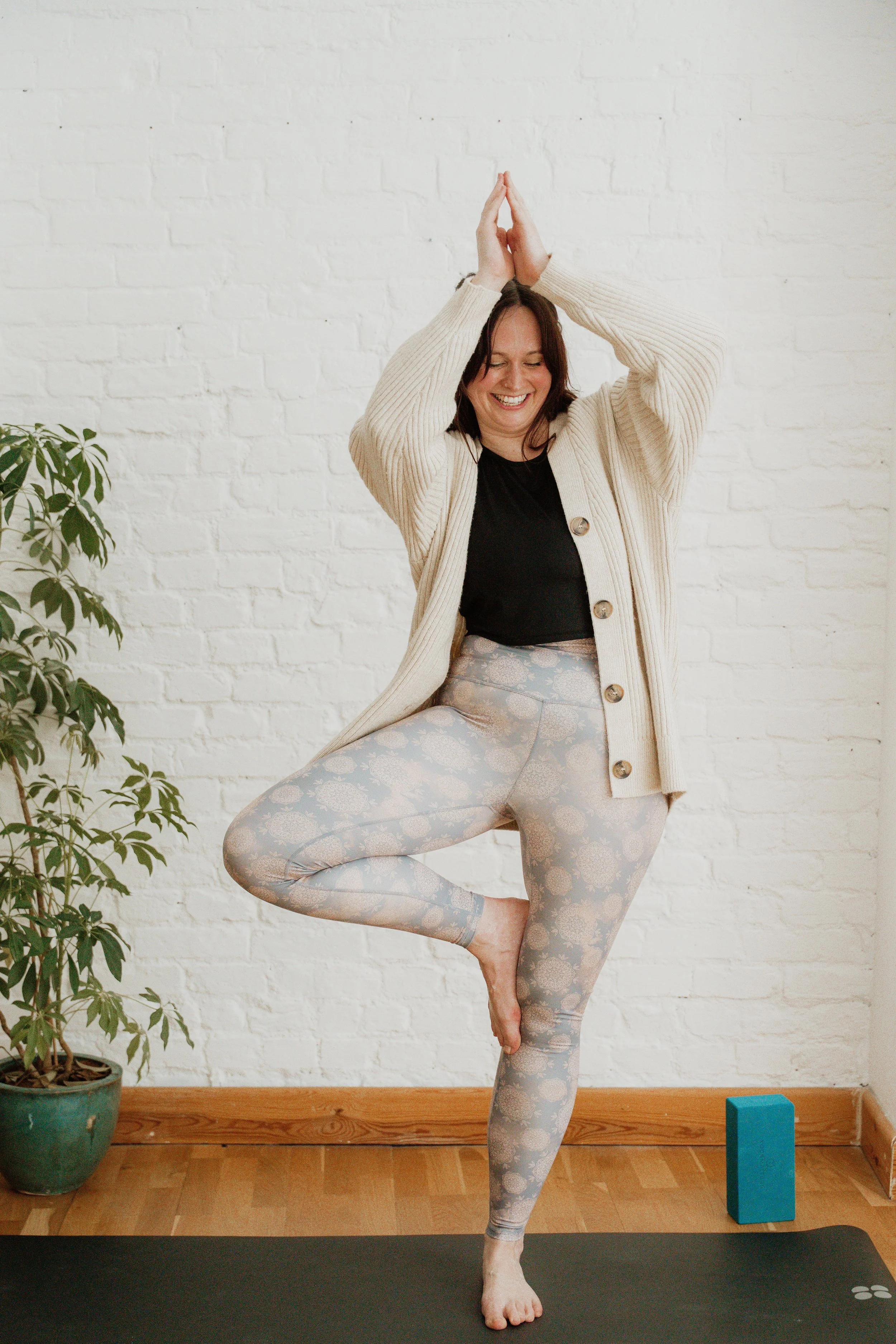 Person practicing yoga in a tree pose, standing on one leg with hands in prayer position above her head. She wears a beige cardigan, black t-shirt, and patterned leggings, in a room with a white brick wall, plant, yoga block, and black yoga mat.