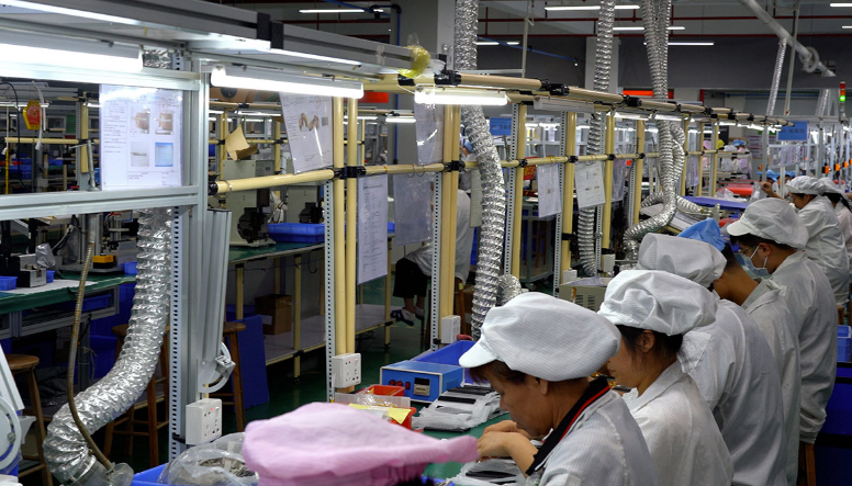 Workers in white uniforms and hats working on assembly line in factory with ventilation ducts and electrical equipment.