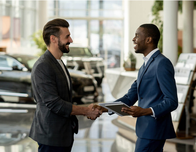 Two men shaking hands inside a car dealership with cars displayed in the background.