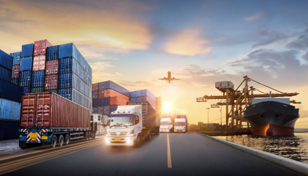 Cargo containers stacked near trucks and ships at a port with cranes, an airplane flying in the sky during sunset.