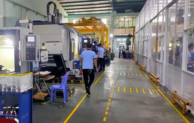 Manufacturing factory with workers in light blue uniforms operating machinery and a safety enclosure on the right side.