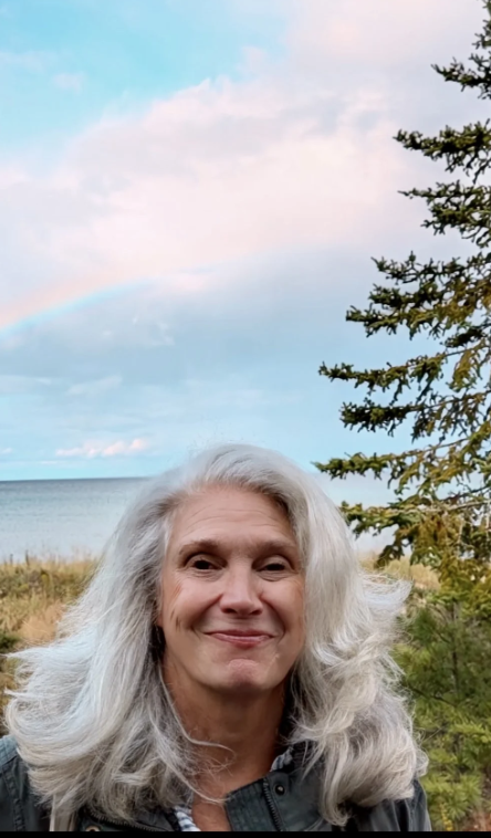 Woman with long gray hair smiling outdoors near a tree, with a rainbow and the ocean in the background.