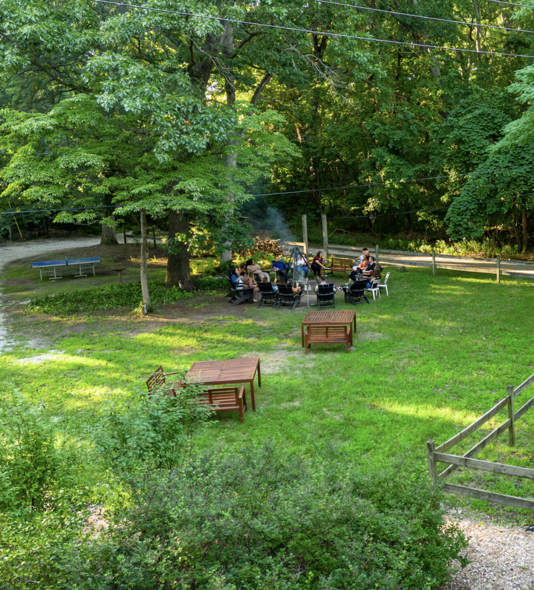 A group of people gathered around a fire pit in a lush green backyard with large trees, wooden benches, and outdoor furniture, enjoying a social gathering in daylight.