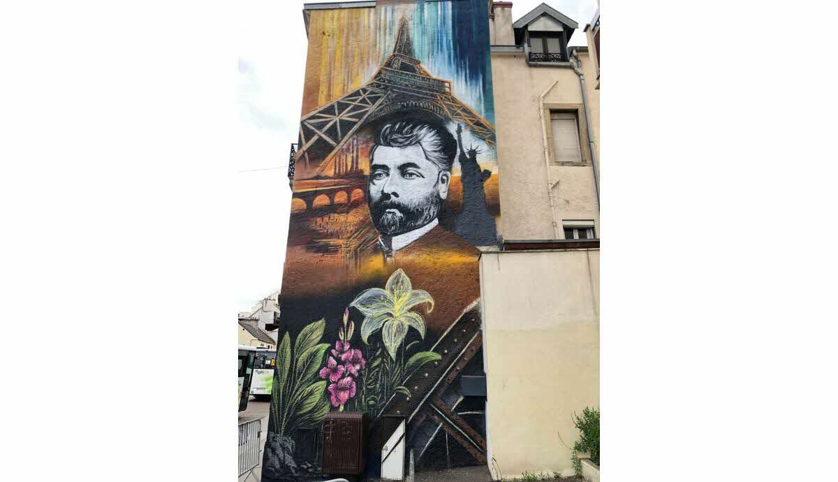 Fresque mural représentant Gustave Eiffel avec la Tour Eiffel en arrière-plan, des fleurs blanches et violettes en bas.