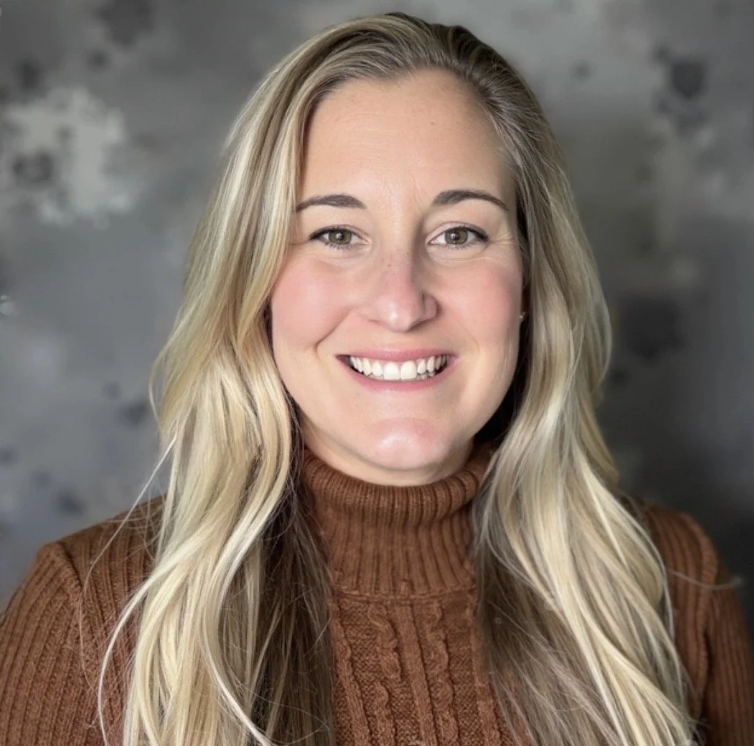 A young woman with long blonde hair, wearing a brown turtleneck sweater, smiling with visible teeth, posing in front of a gray, textured background.