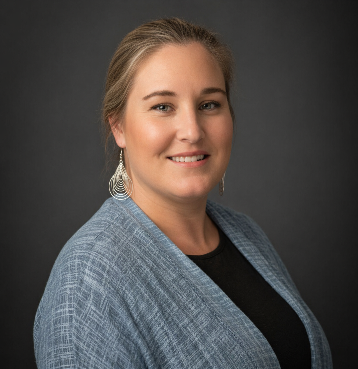 Portrait of a woman with blonde hair, wearing teardrop earrings and a blue blazer, smiling against a dark gray background.