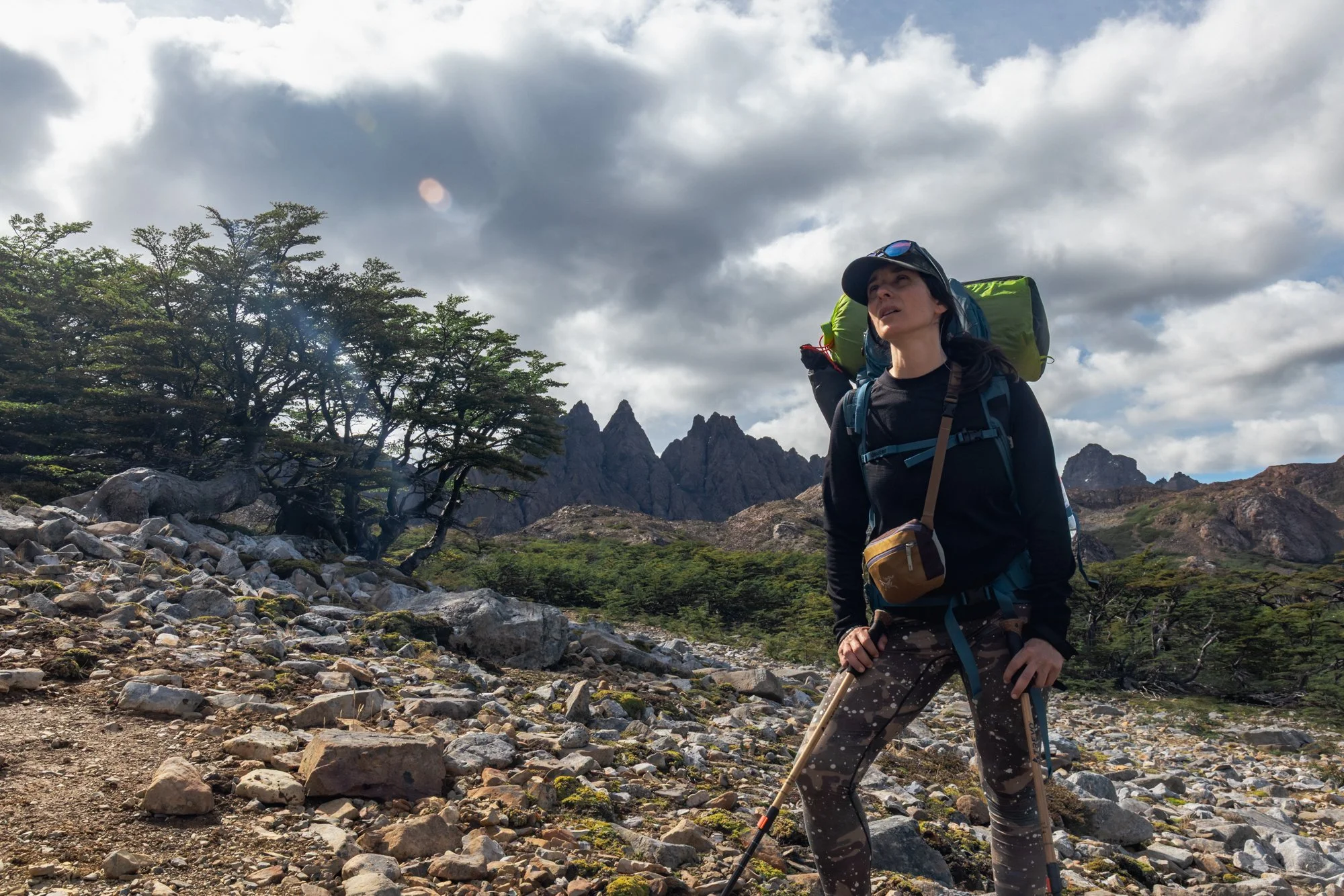 A woman hiking in a mountainous landscape with rocky terrain, green trees, and mountains in the background, under a cloudy sky.