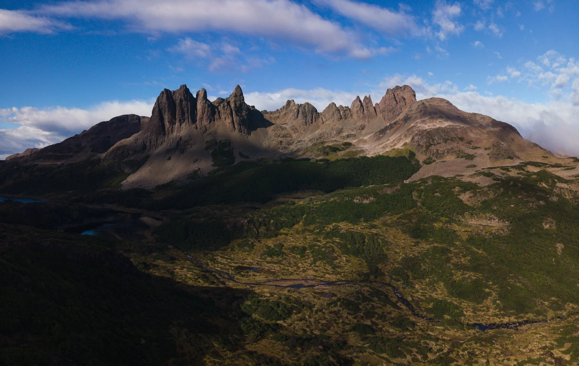 A mountain range with rugged peaks under a partly cloudy sky, with green vegetation at the base.