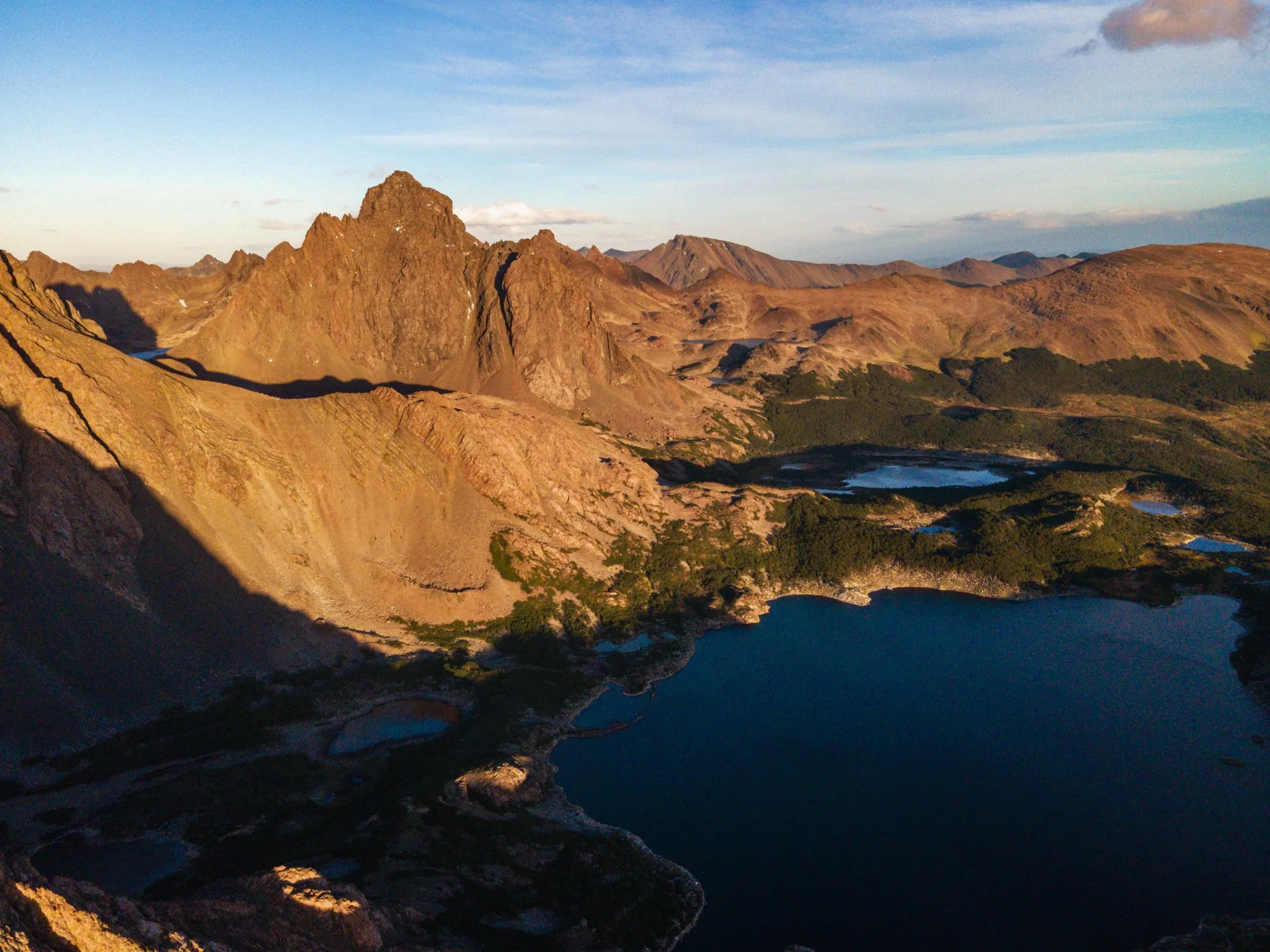 Photograph of a mountain range with rugged peaks, a lake, and a partly cloudy sky during daytime.