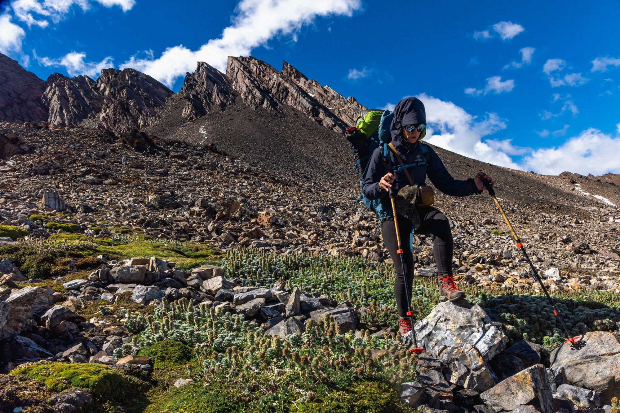A person hiking in a rocky mountain terrain under a blue sky with clouds, wearing a dark jacket, sunglasses, and carrying a green backpack with hiking poles.