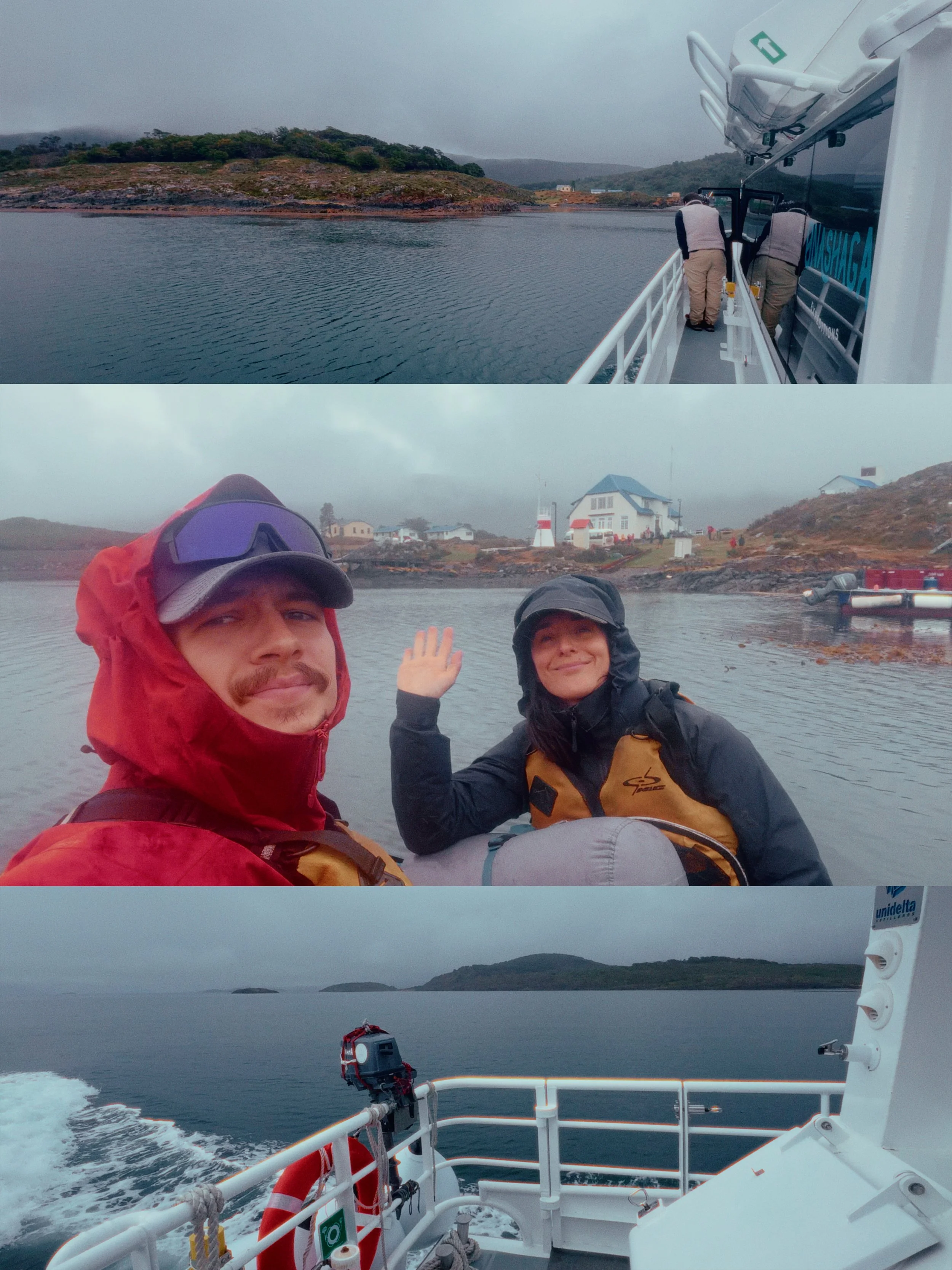 View from a boat showing two people in rain gear smiling and waving, with a coastline featuring houses and trees in the background, overcast sky and calm waters.
