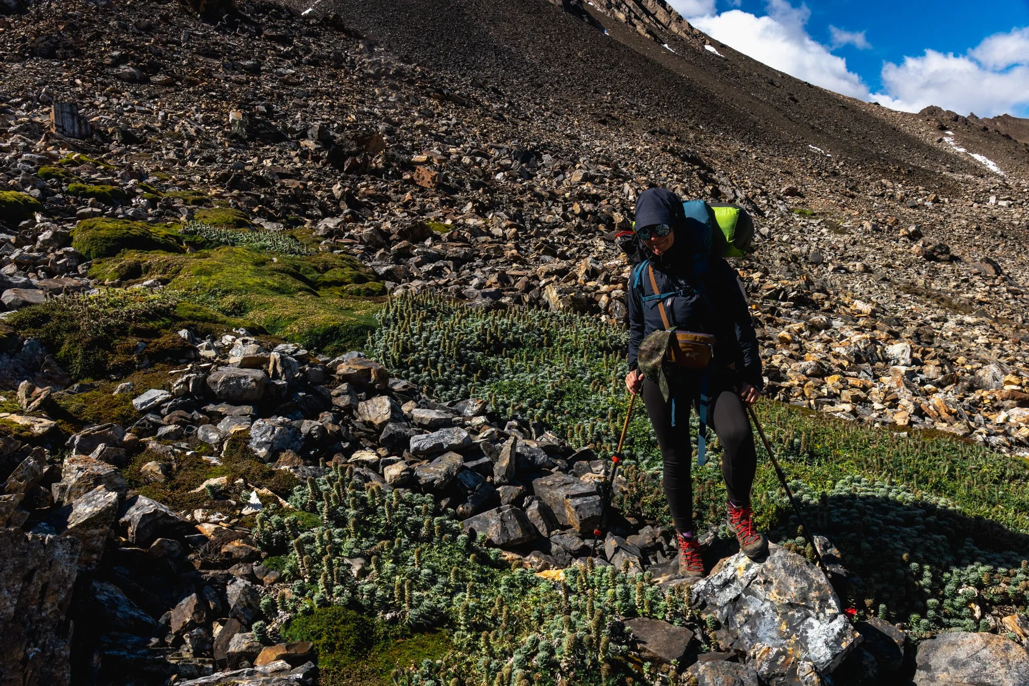 Hiker in dark outdoor clothing and red hiking boots climbing on steep rocky terrain with moss and cactus, under a partly cloudy sky.